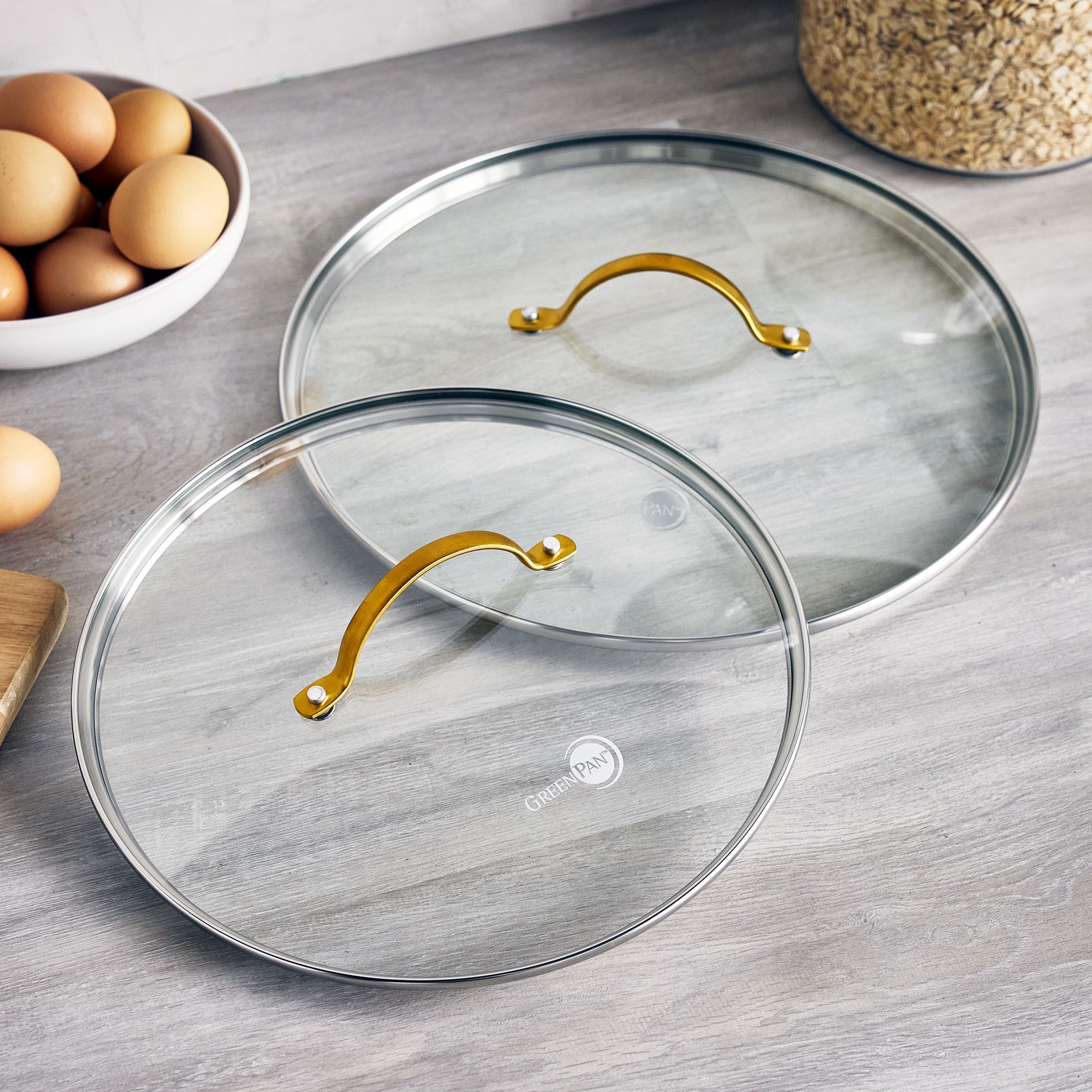 Two glass pan lids with gold handles on a wooden surface, next to brown eggs and a bowl of oats.