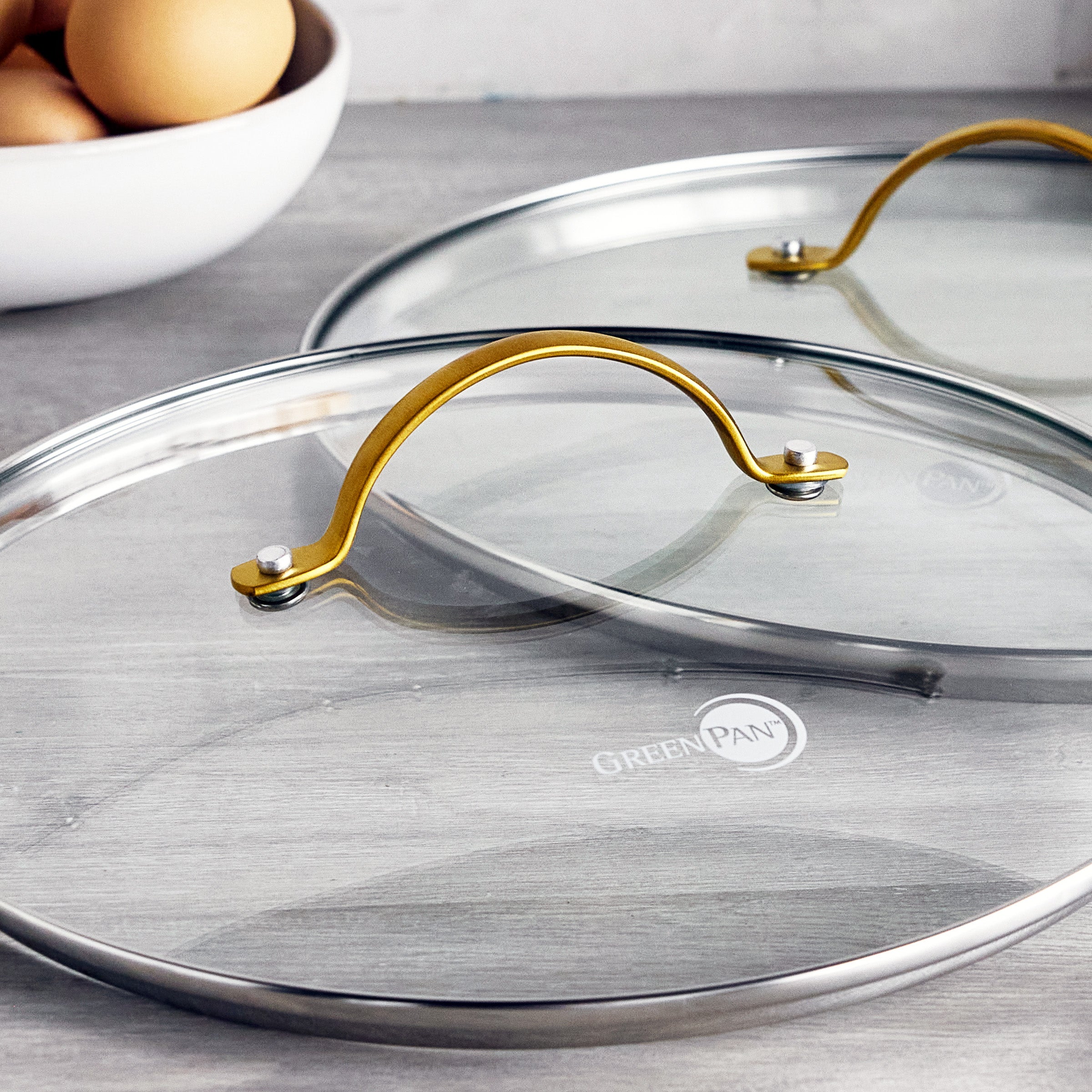Set of two glass pot lids with gold handles on a gray surface; white bowl of brown eggs in the background.