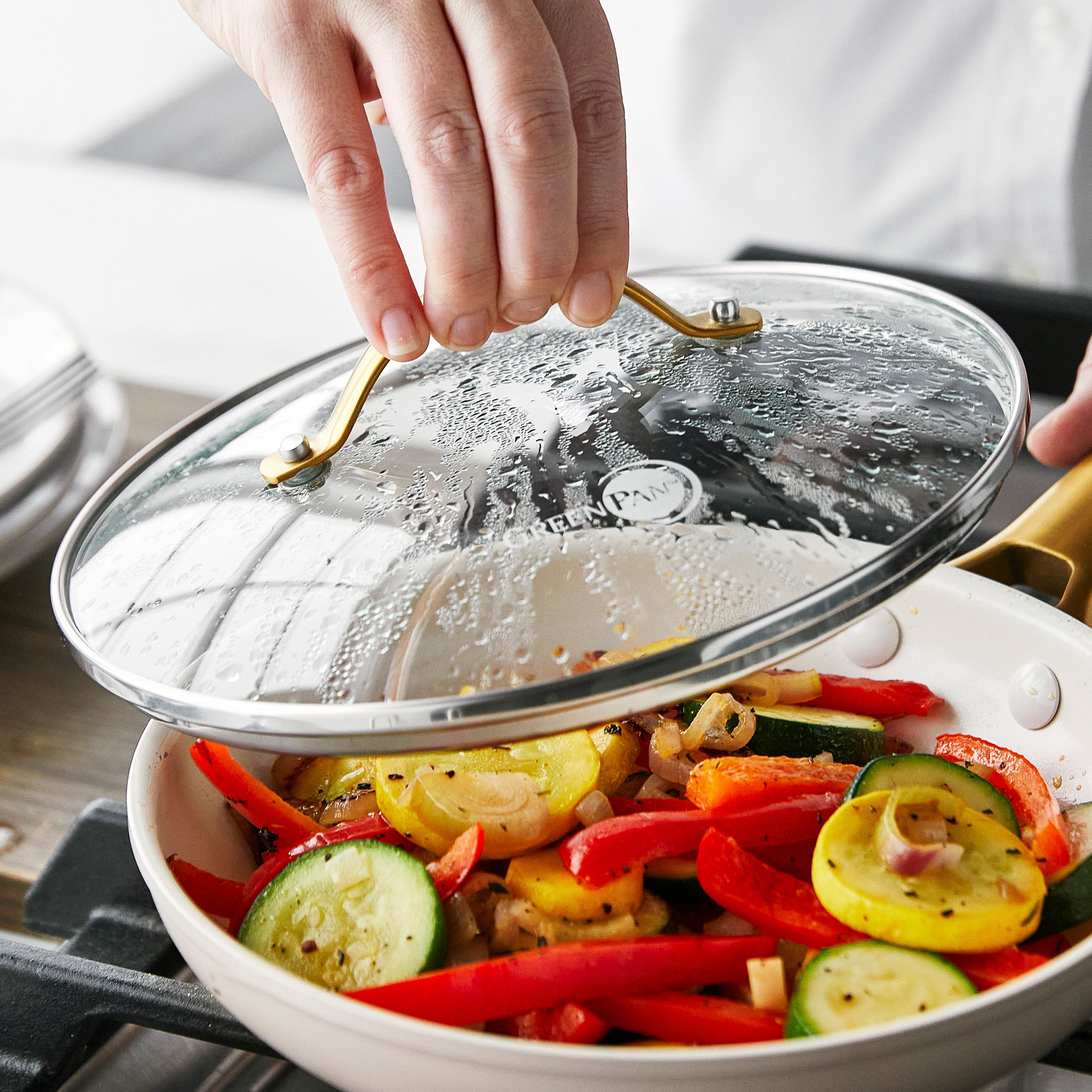 Hand lifting a glass GreenPan lid from a pan with sautéed zucchini, squash, and red bell pepper.