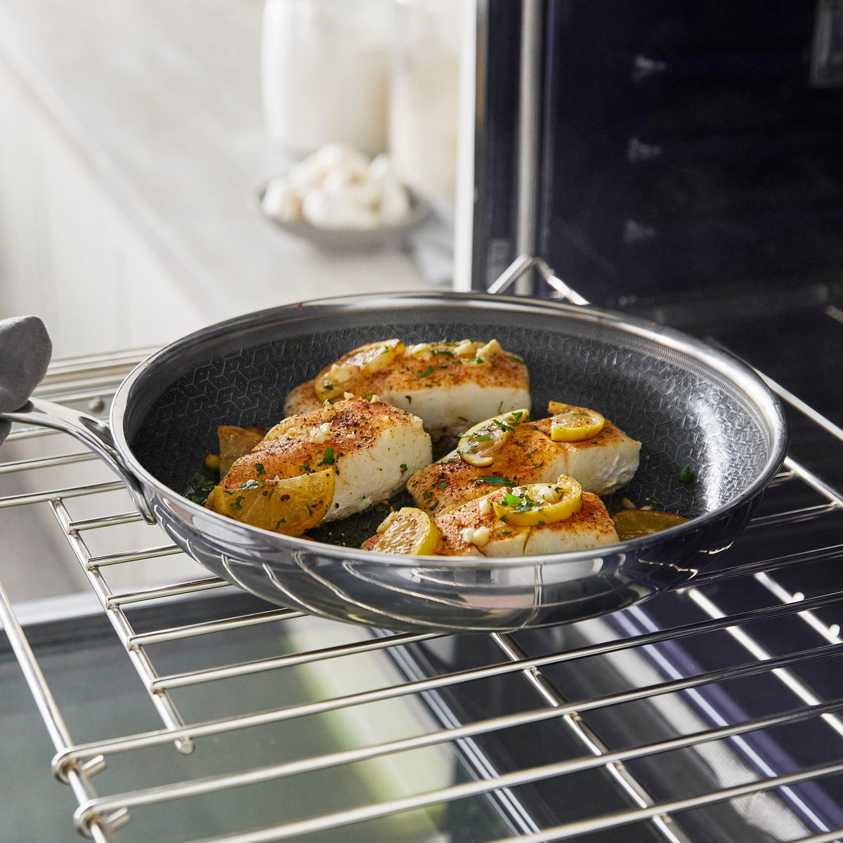 Seared fish fillets and lemon slices in a dark, textured Spectra Diamond Thermobond hybrid ceramic nonstick frypan on an oven rack.