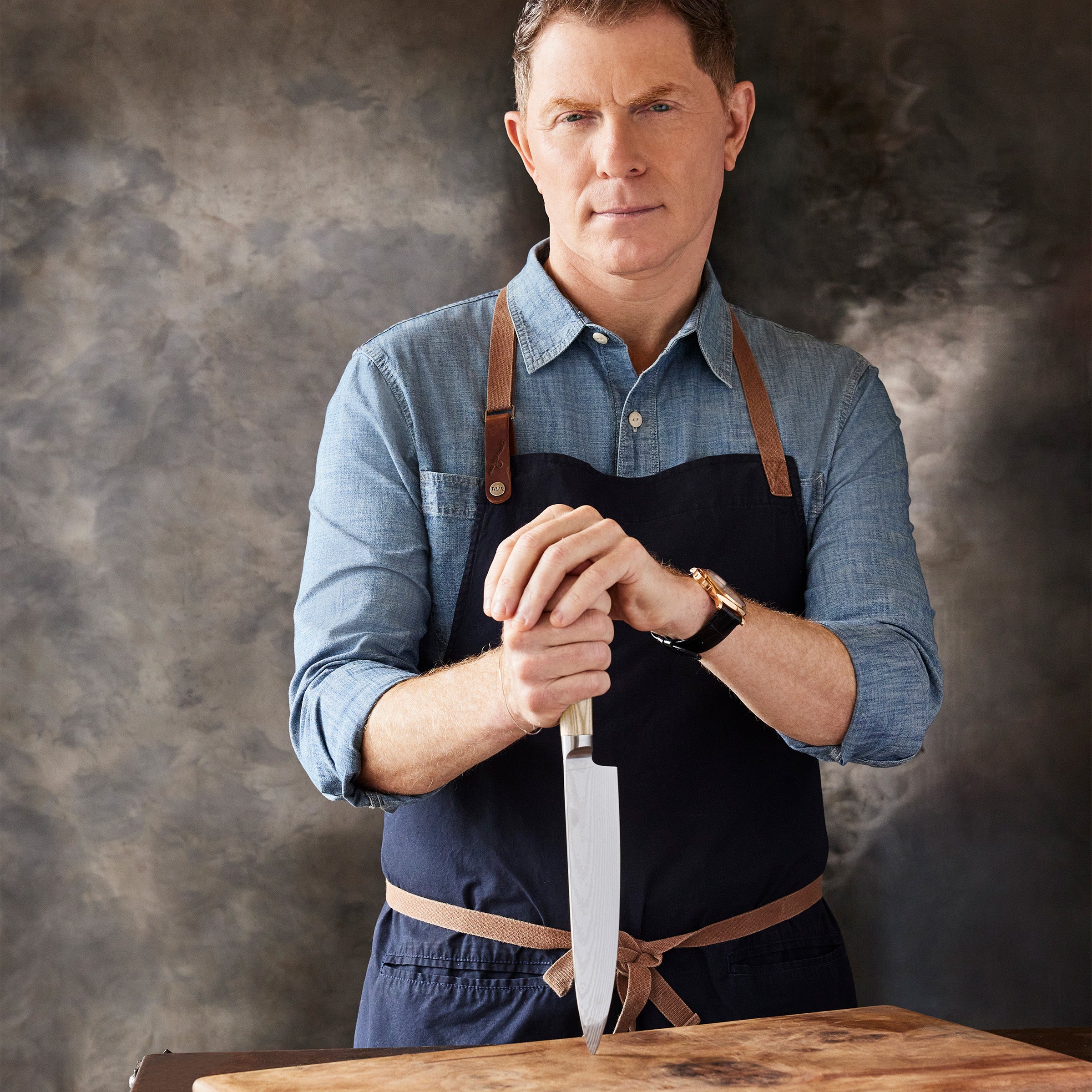 Bobby Flay in a blue shirt and apron holding a knife on a wooden cutting board against a textured gray background