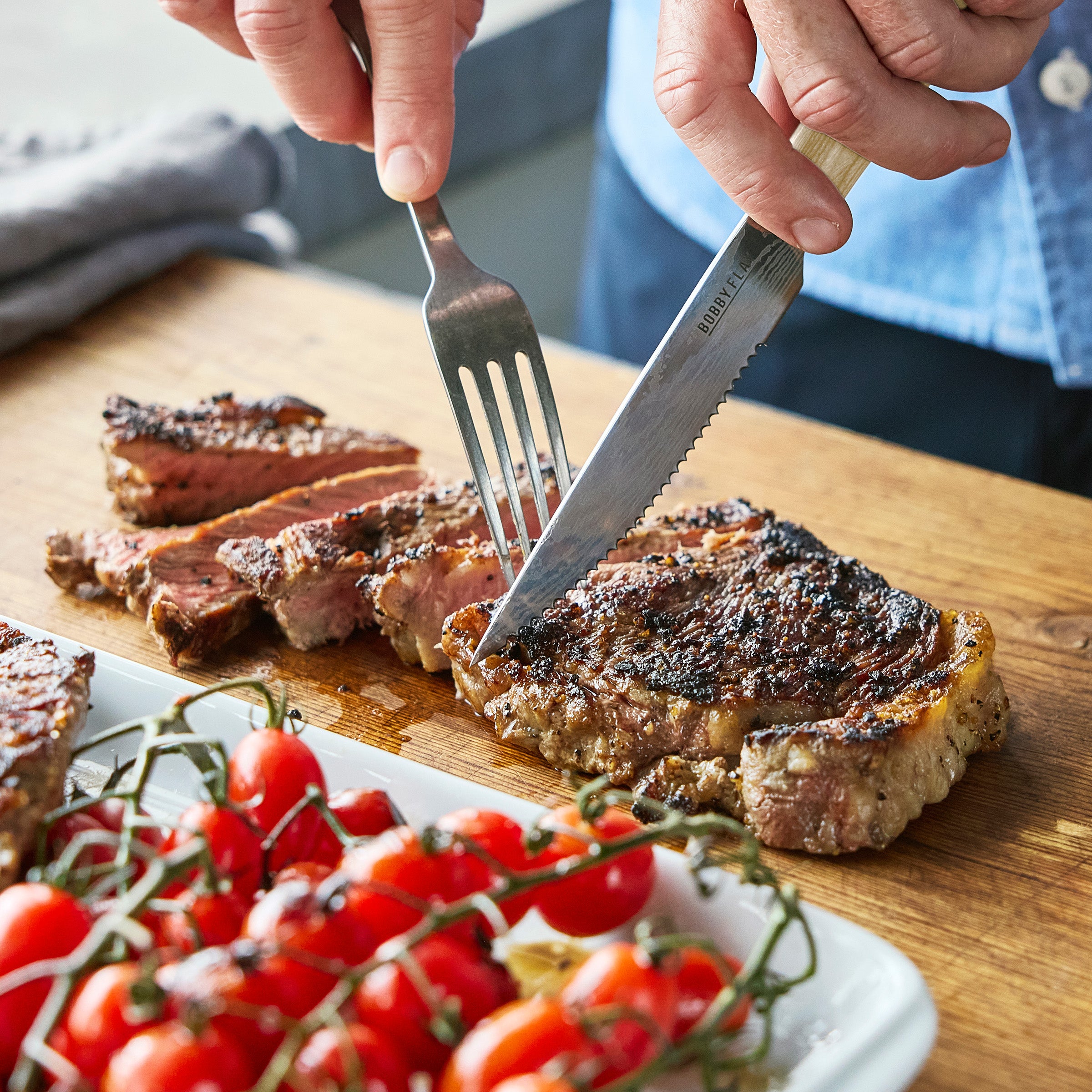 Person cutting a steak with a fork and knife on a wooden cutting board, with cherry tomatoes in the foreground.