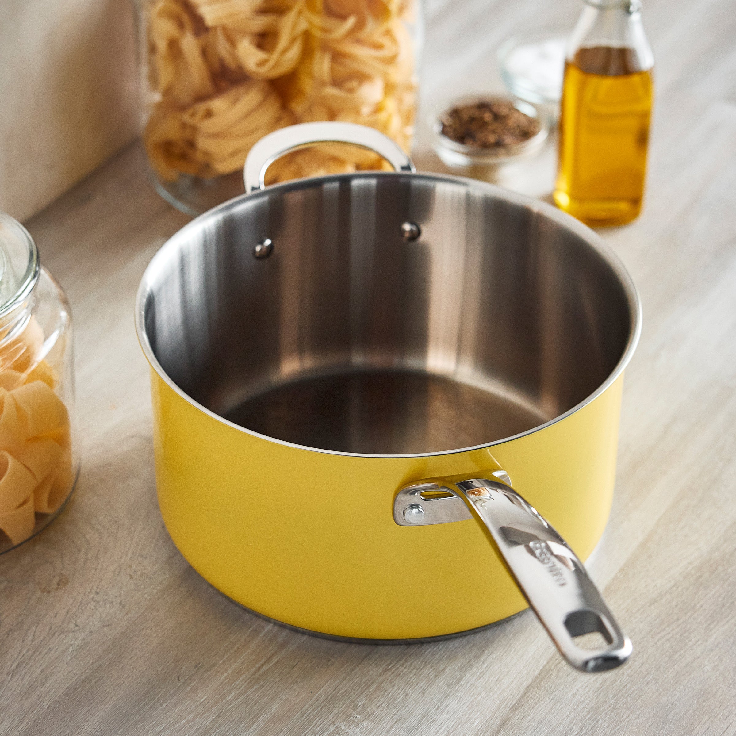 Yellow popcorn maker on a wooden surface with glass jars of pasta, olive oil bottle, pepper grinder, and a yellow saucepan nearby.