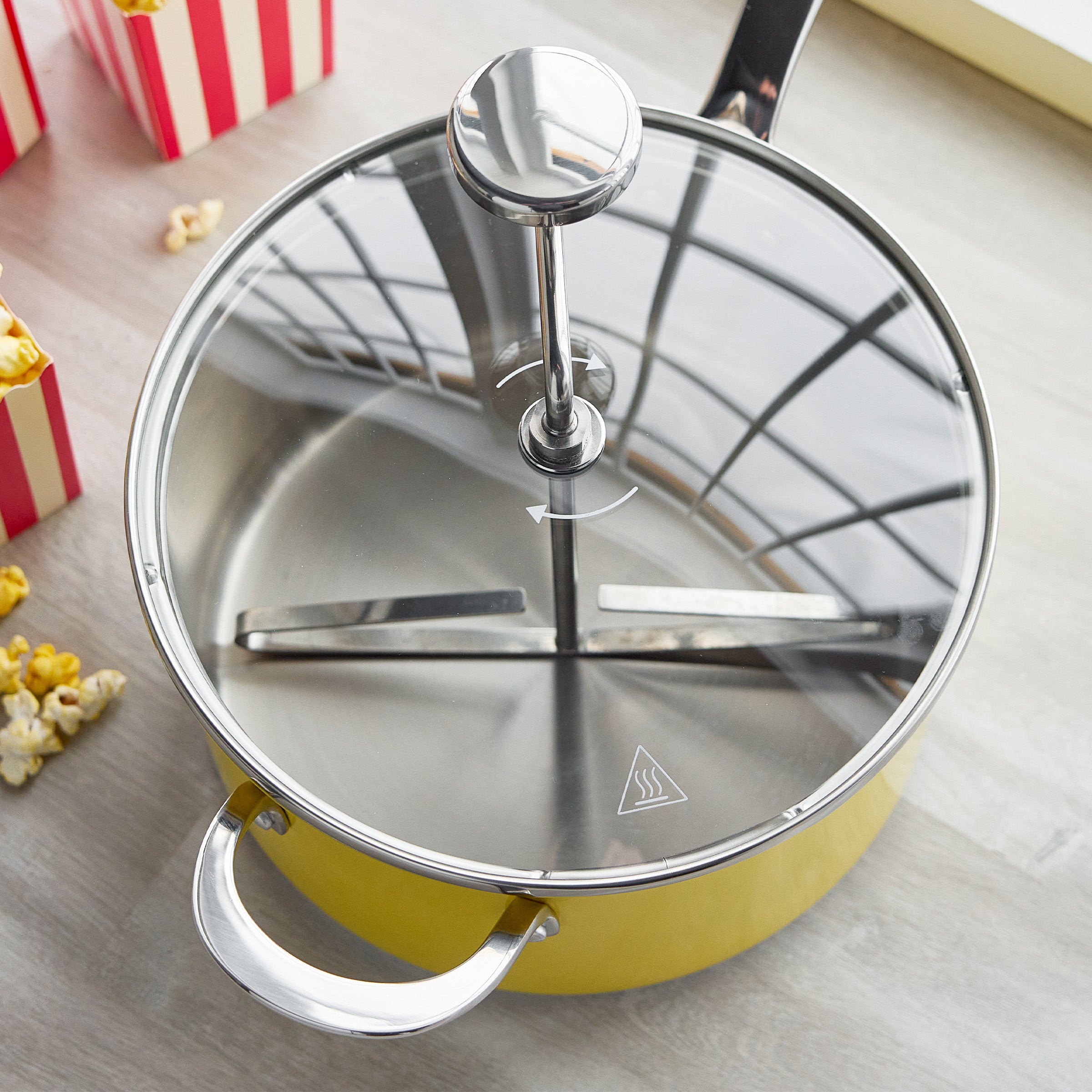 Yellow popcorn maker with stainless steel handle and clear lid next to popped popcorn and striped boxes on a light background.