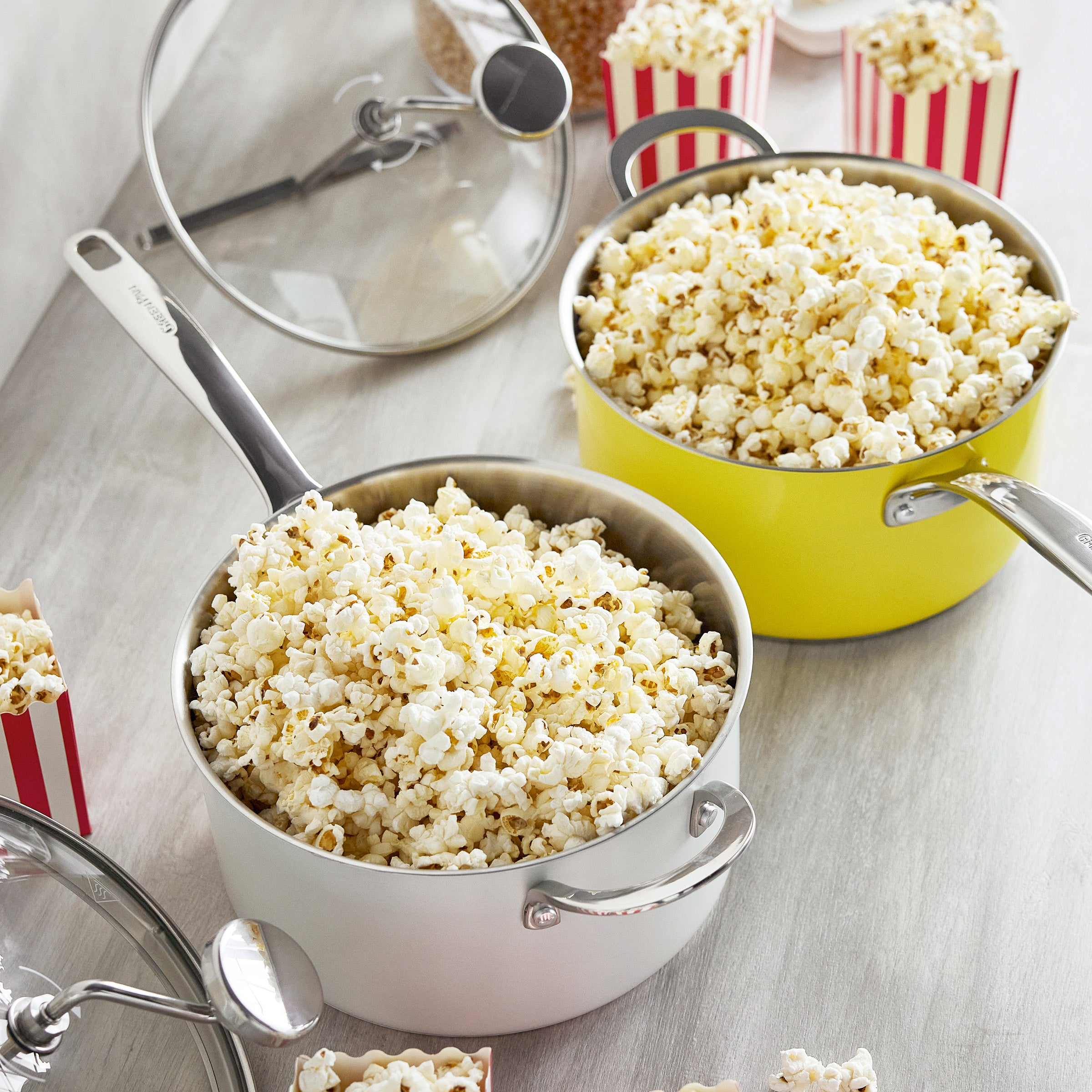 Two yellow popcorn makers on a table with popped popcorn, small boxes, and clear glass lids nearby.