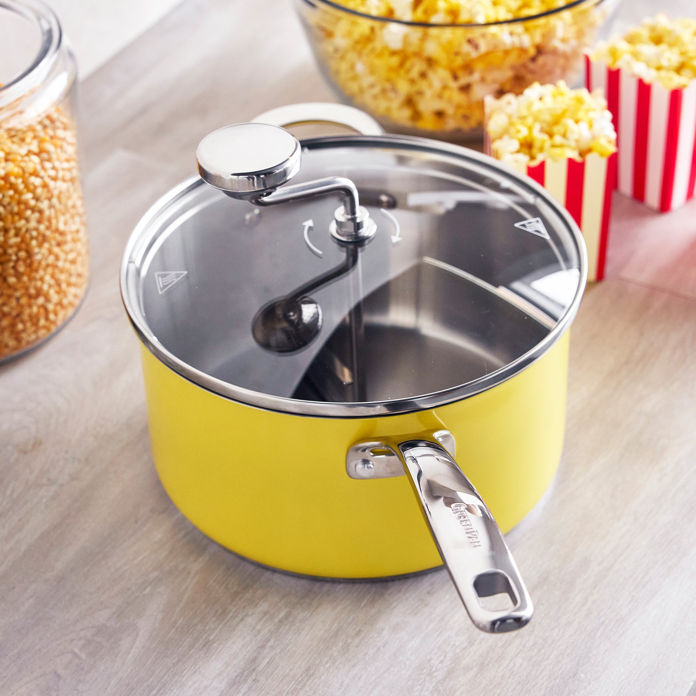 Yellow popcorn maker with glass lid on a table, surrounded by popped popcorn, kernels, and red-striped boxes.