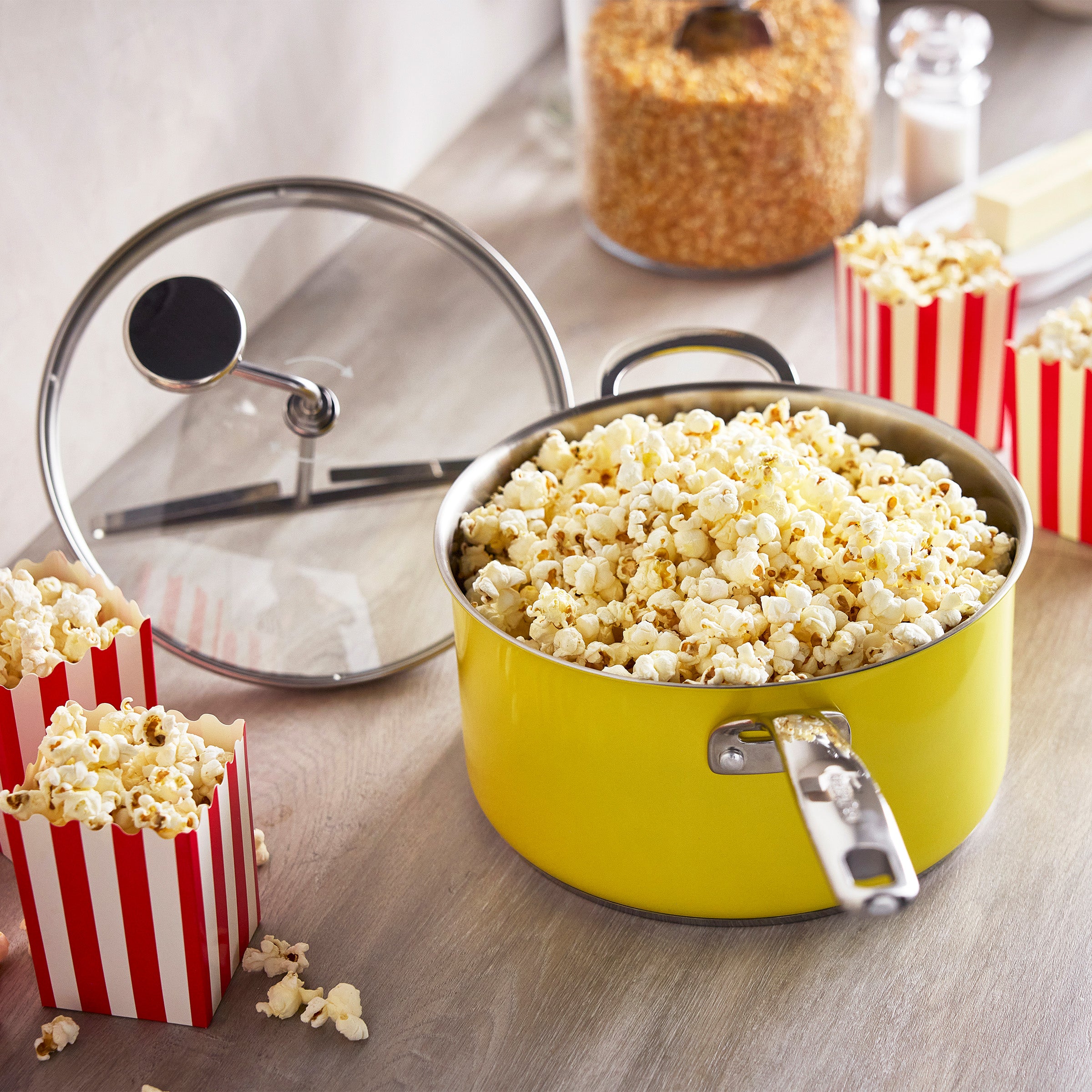Yellow popcorn maker with glass lid on table, surrounded by popcorn and striped serving boxes.