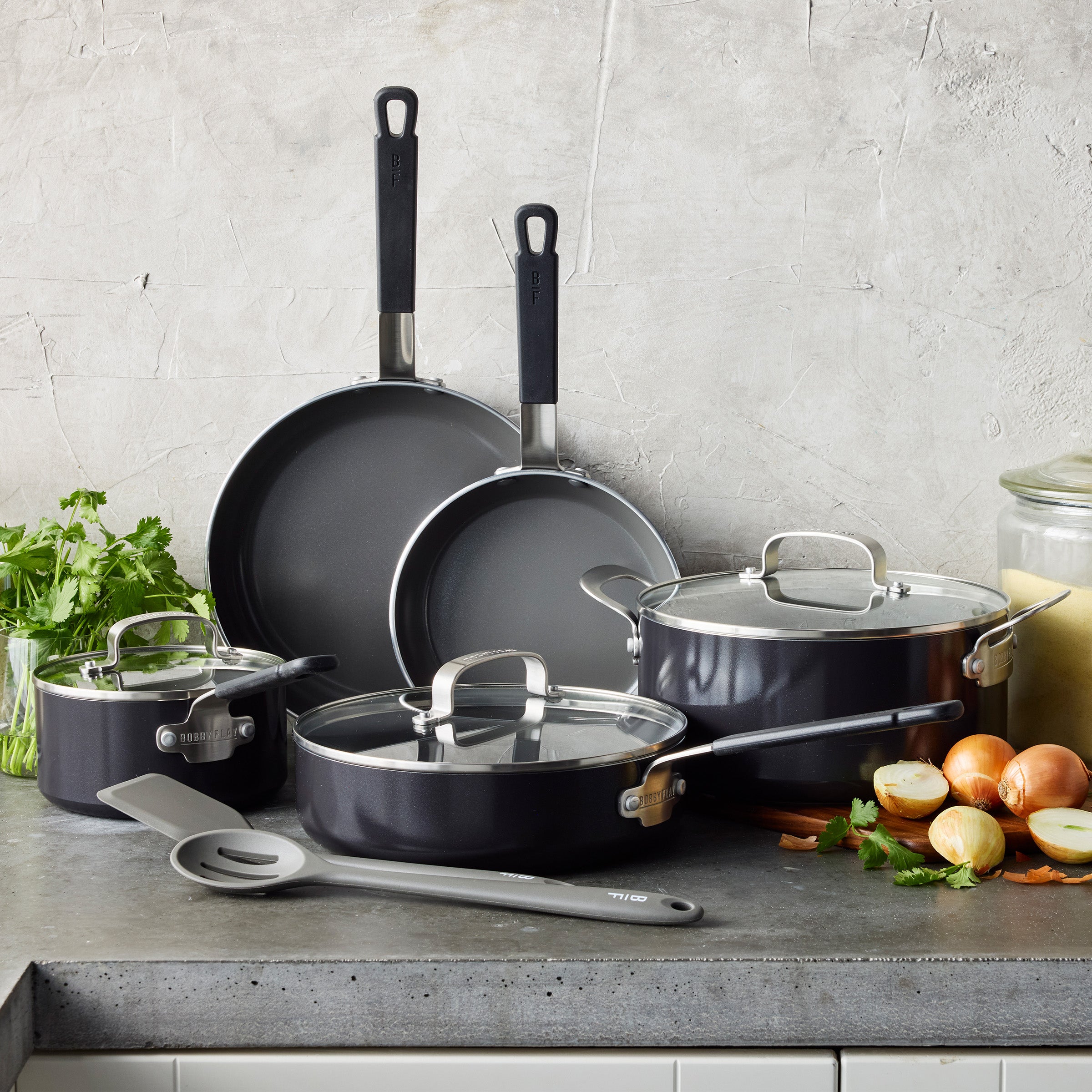 10-piece black cookware set with lids on a gray counter, surrounded by herbs, red onions, and kitchen utensil.