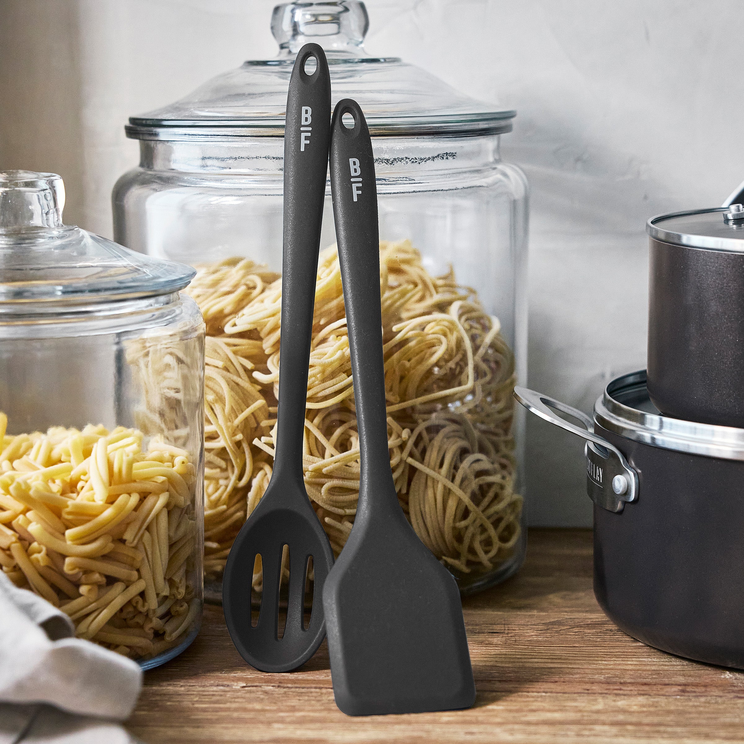 Black utensils rest against pasta jars near a black GreenPan ceramic nonstick cookware set on a wooden countertop.