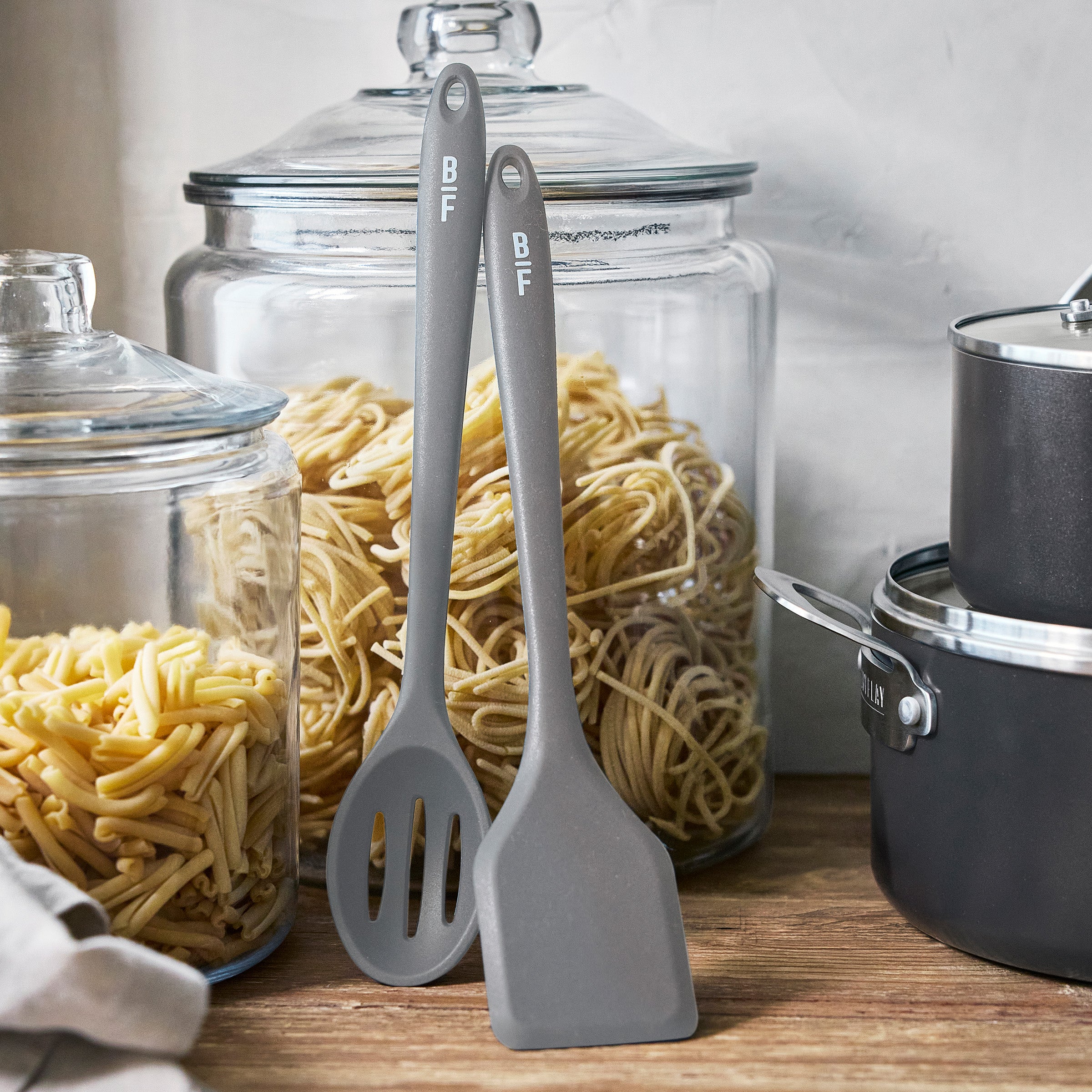 Graphite ceramic nonstick cookware set with two gray utensils and glass jars of pasta on a kitchen counter.