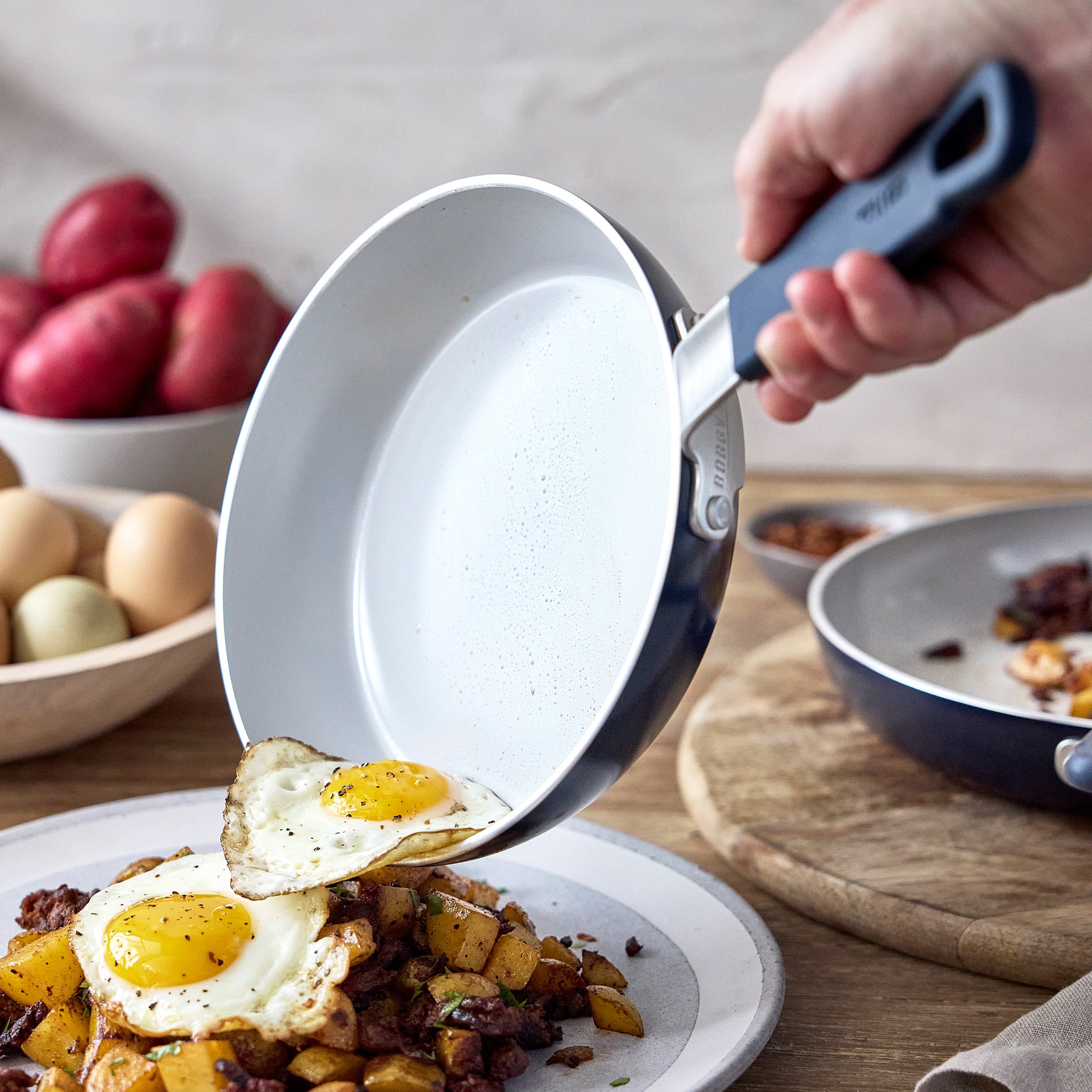 Hand sliding eggs from a Bobby Flay by GreenPan ceramic nonstick pan onto potatoes and meat on a plate.