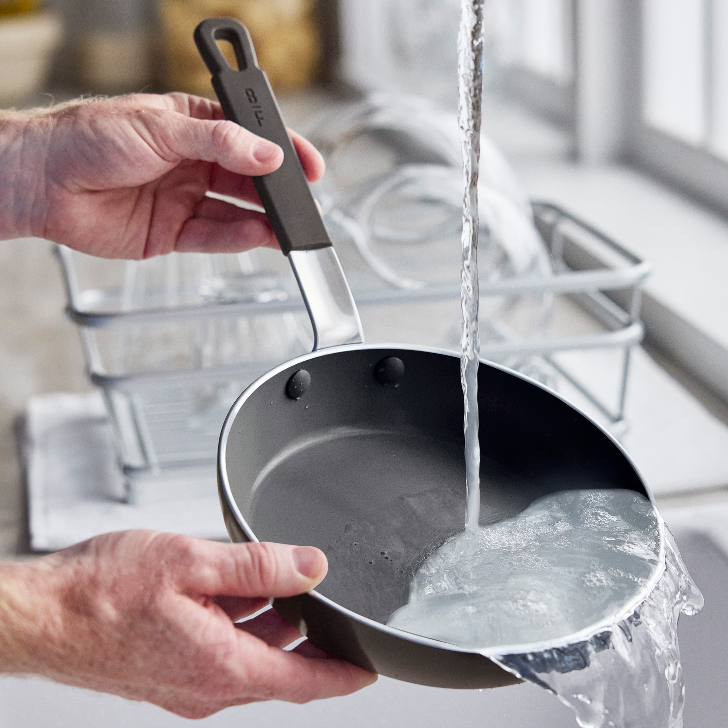 Ceramic nonstick pan being washed under running water in a bright kitchen, dish rack nearby.
