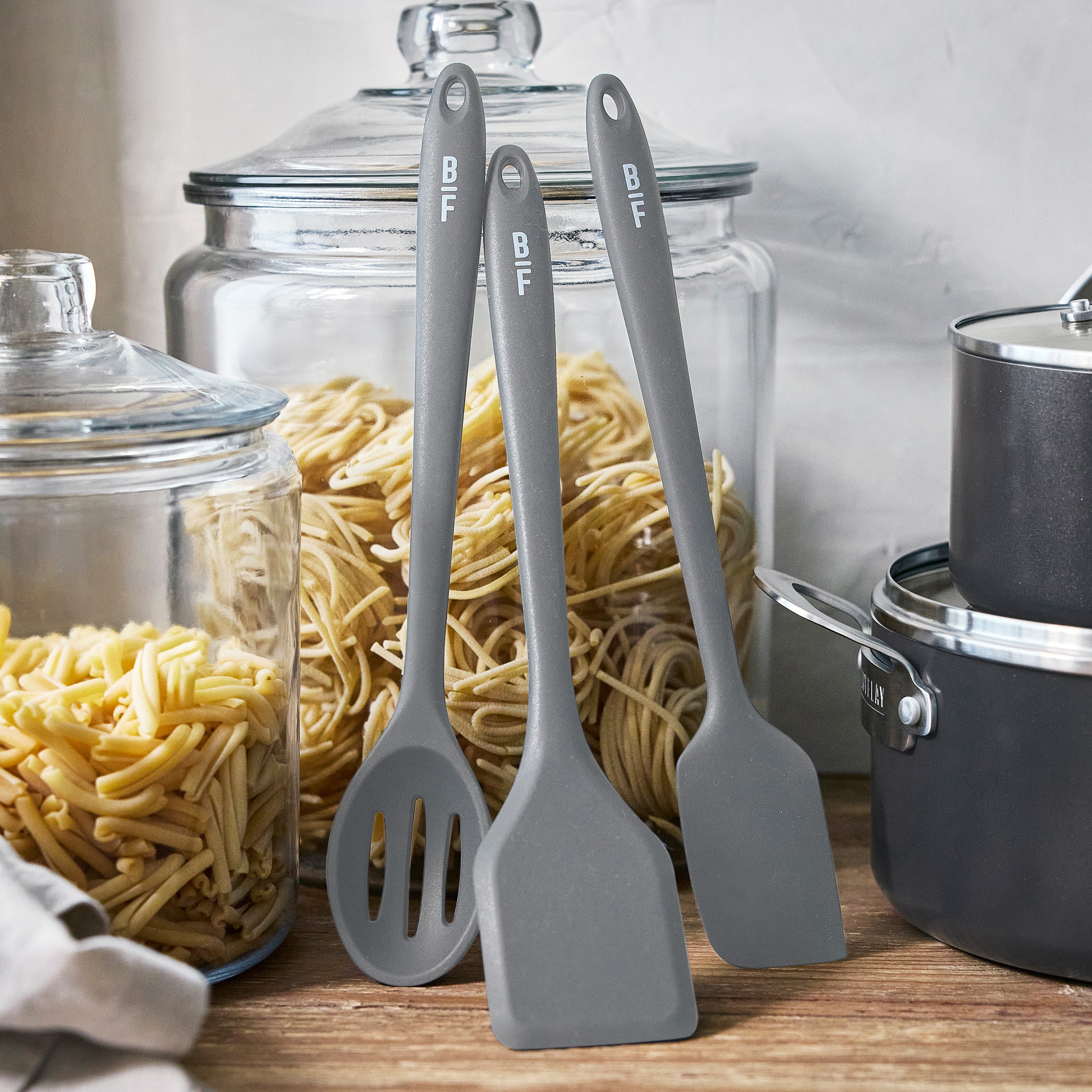 Gray kitchen utensils and jars of pasta beside a Bobby Flay by GreenPan cookware set on a kitchen counter.