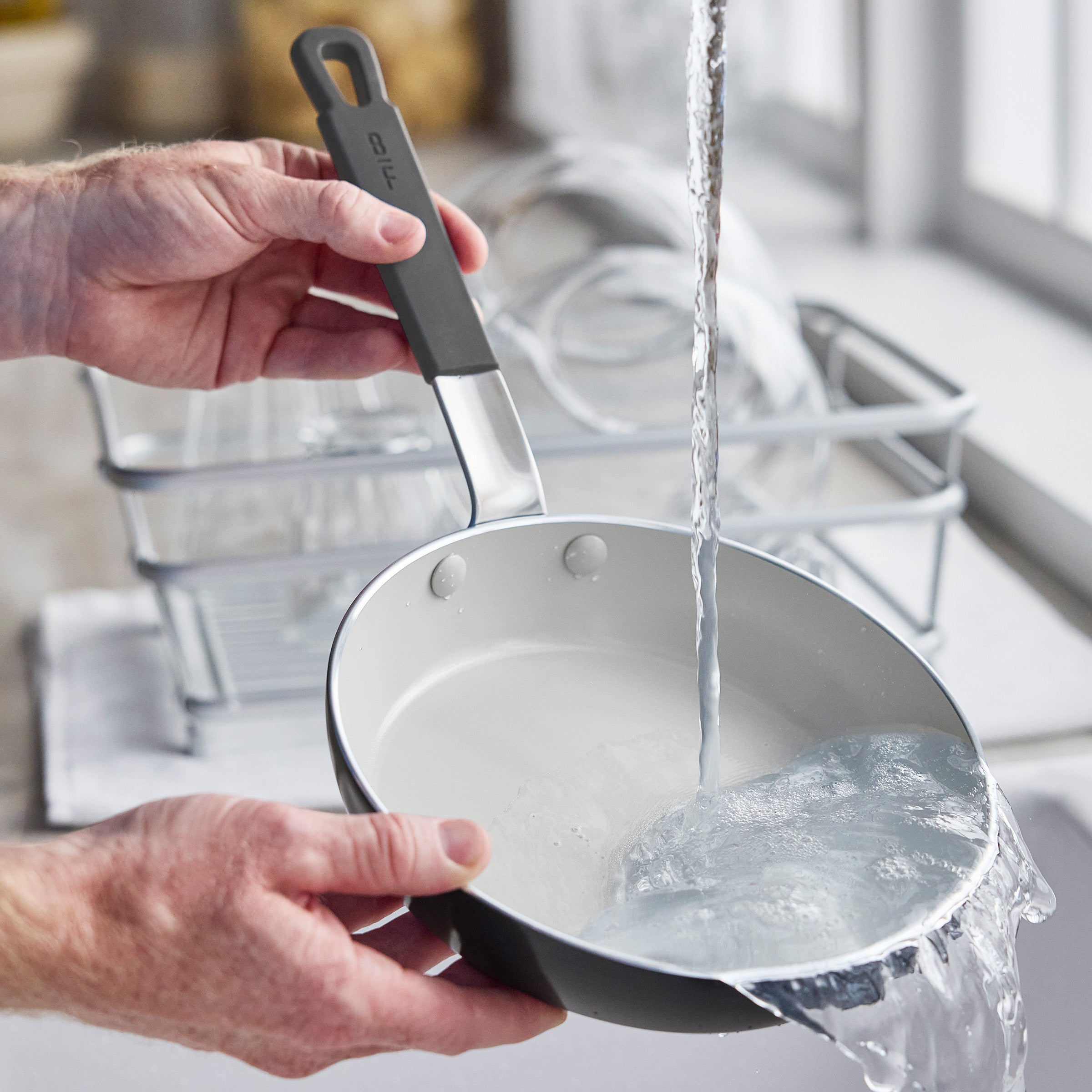 Washing a GreenPan ceramic nonstick pan at the sink, with a dish rack and window in the background.