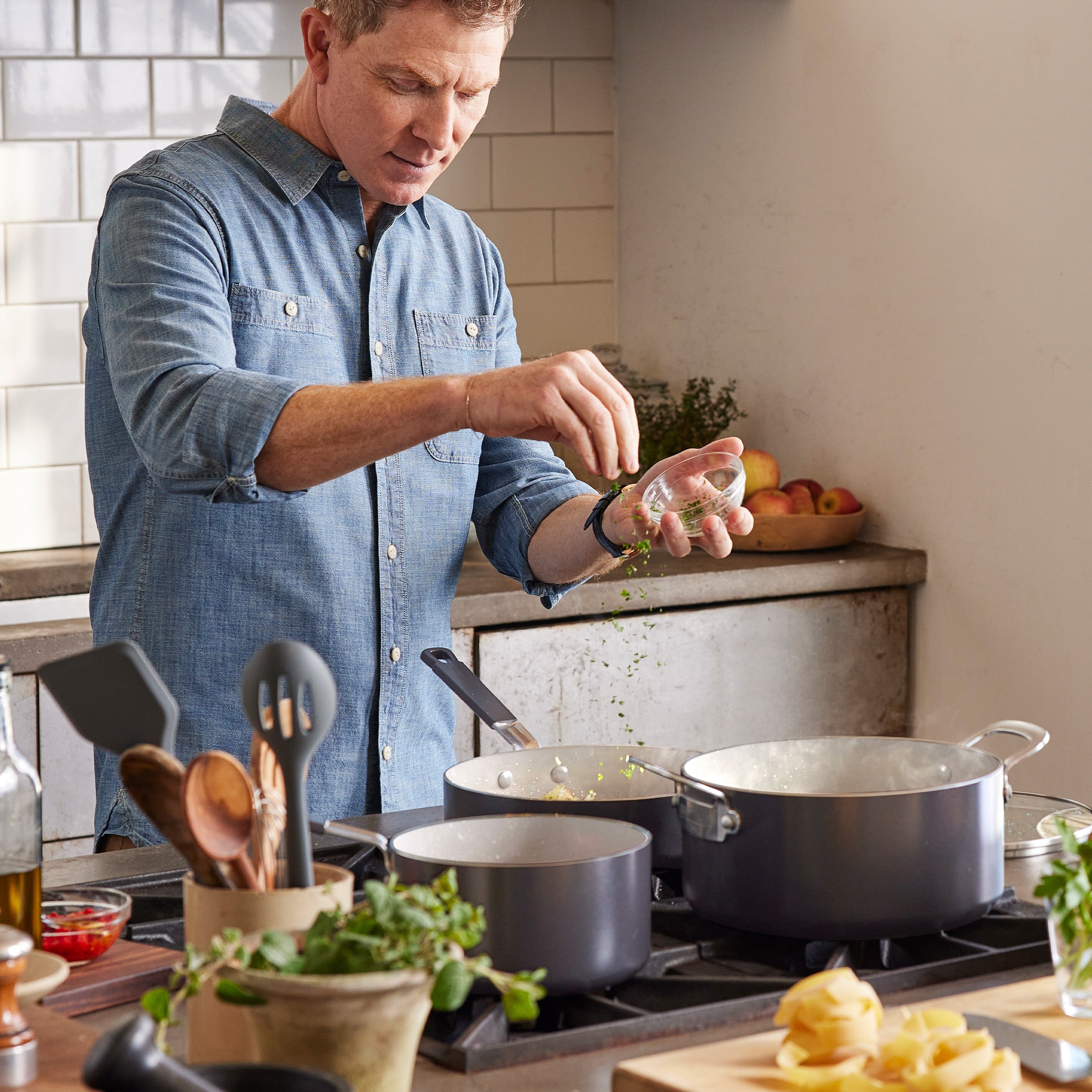 Man sprinkles herbs into pot on stove, using Bobby Flay by GreenPan 16-piece cookware set, surrounded by fresh ingredients.