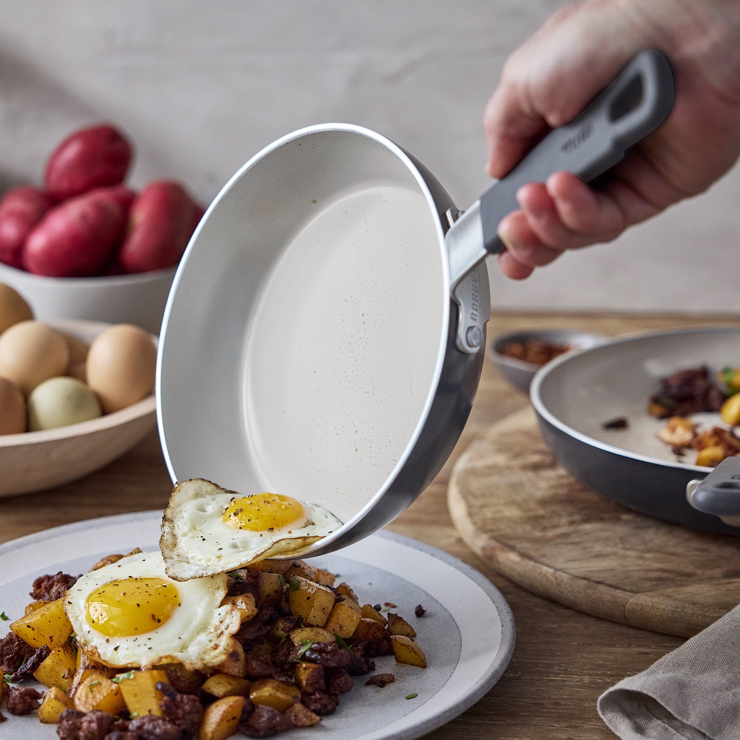 Hand slides two fried eggs from a GreenPan nonstick pan onto potatoes and meat; eggs and potatoes visible in the background.