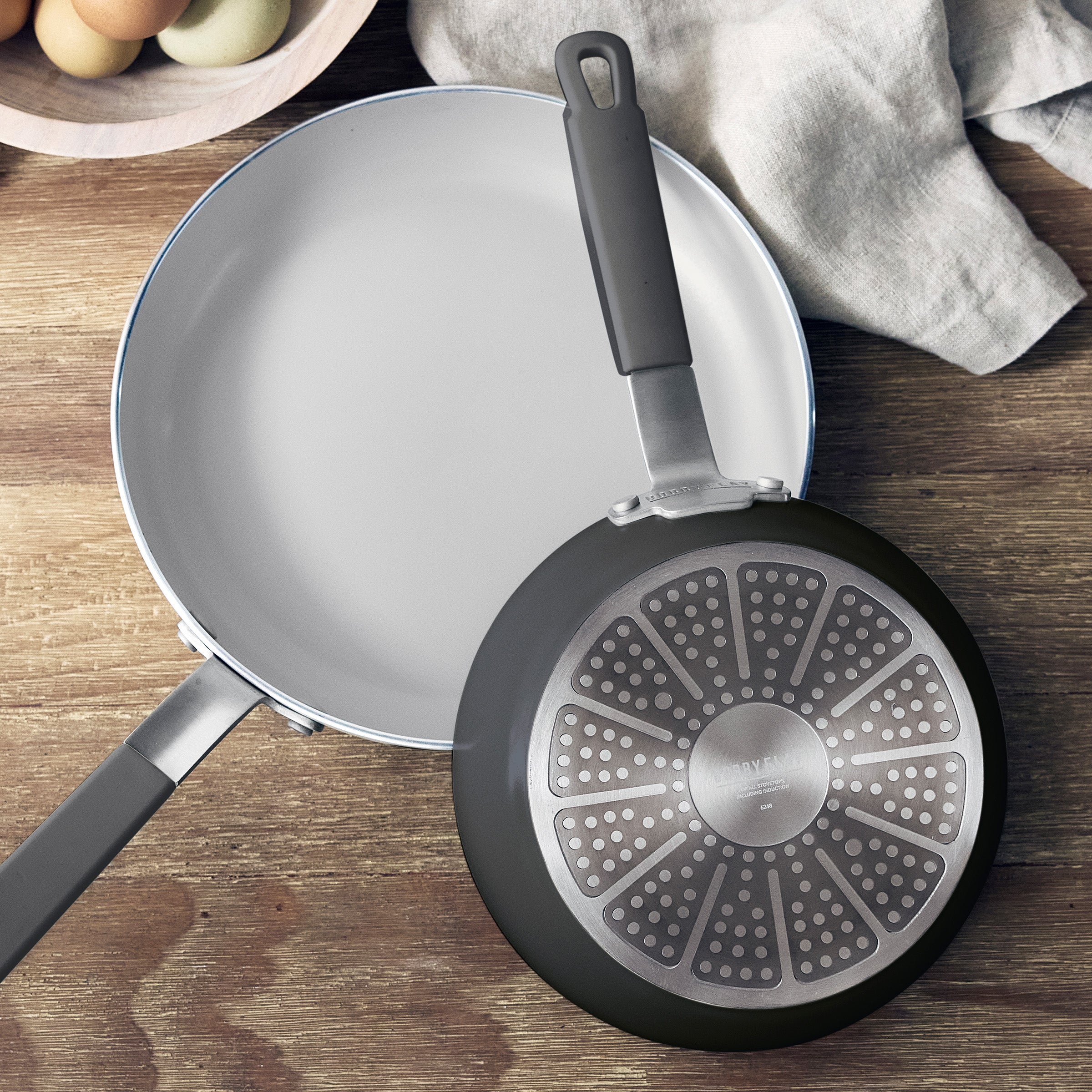 Two ceramic nonstick frying pans, one upright and one upside down, on a wooden surface with eggs beside them.