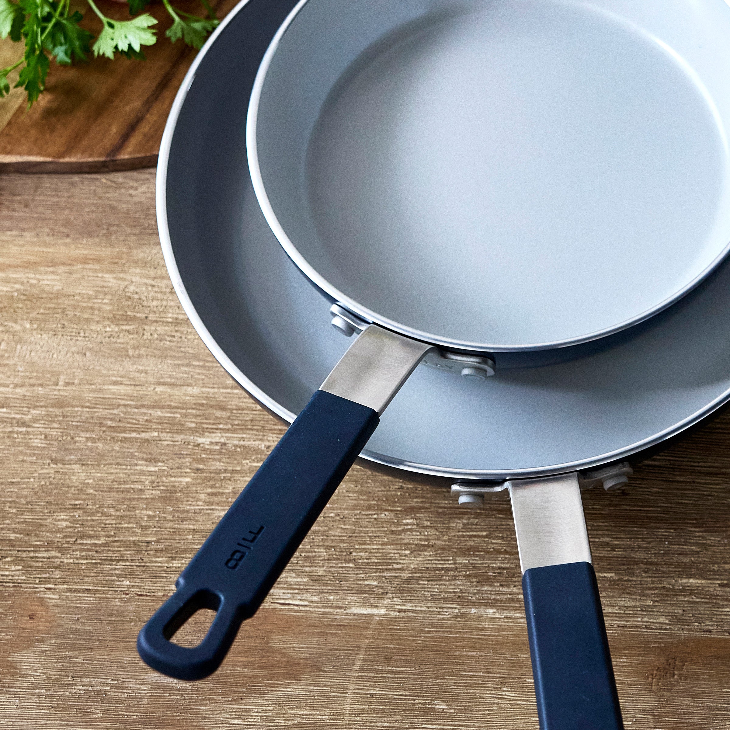 Two stacked blue Bobby Flay by GreenPan pans with black handles on a wood surface, herbs in the corner.