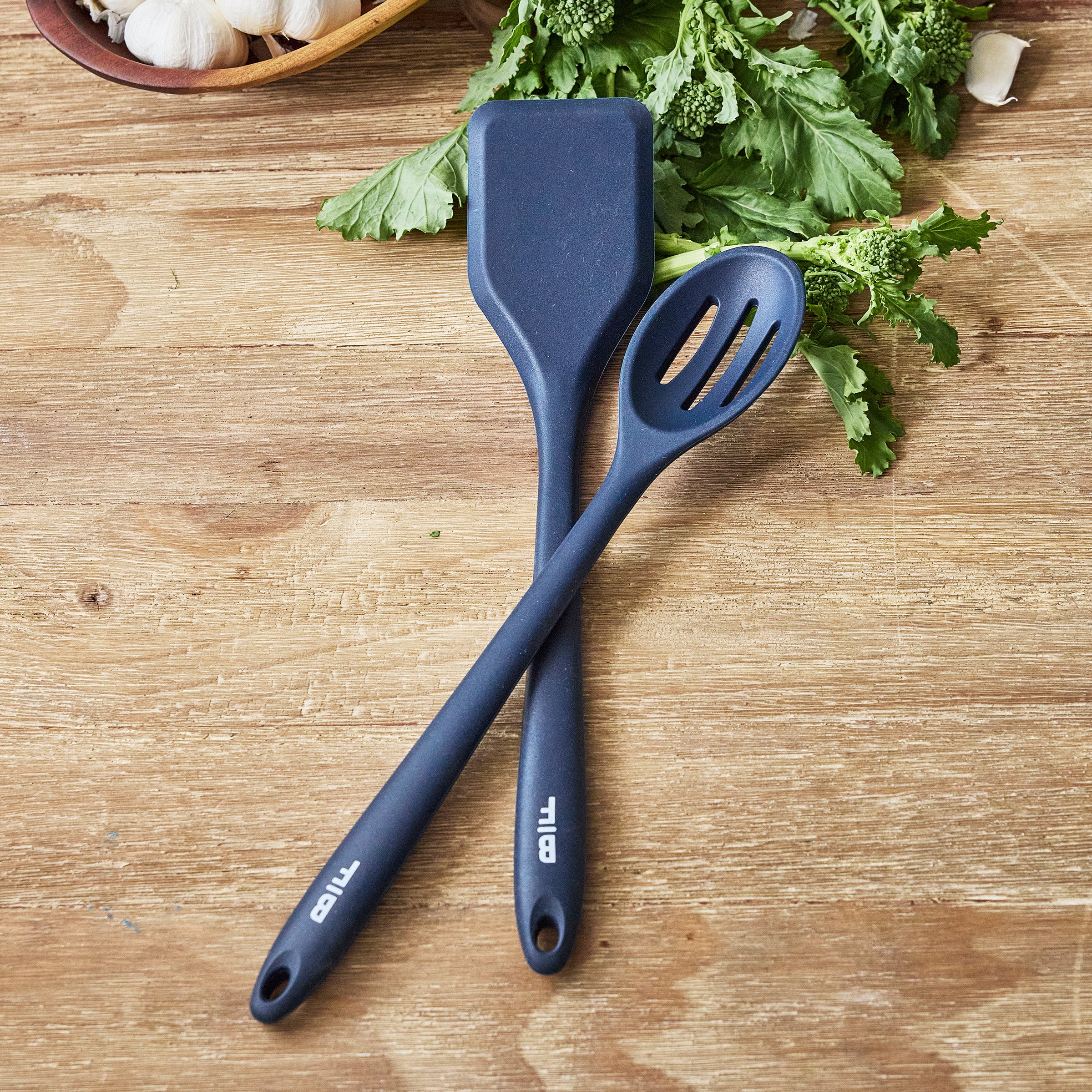 Two Oxford Blue Bobby Flay by GreenPan utensils on a wooden table with leafy greens blurred in the background.
