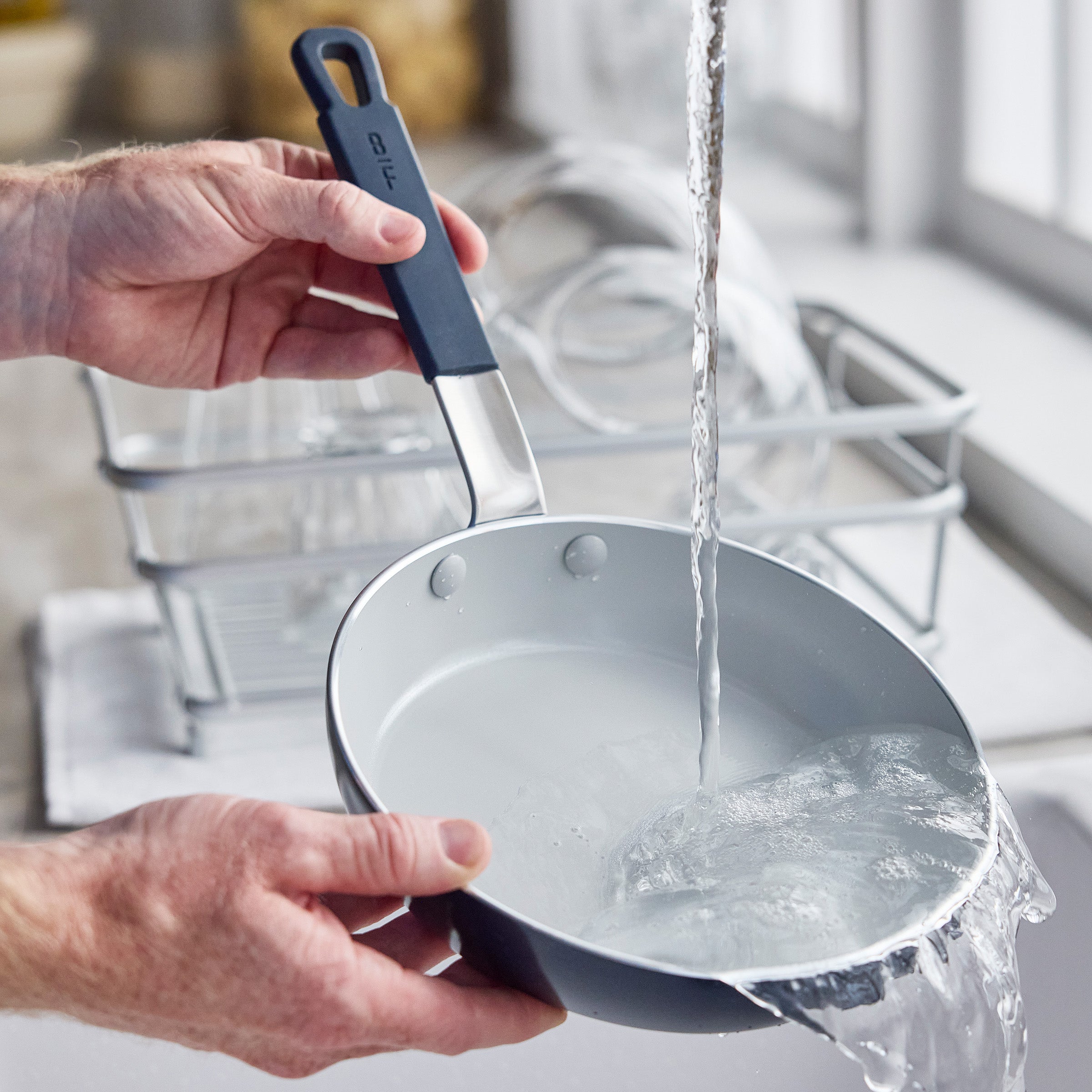 Blue nonstick pan being washed under running water in a bright kitchen, drying rack nearby.