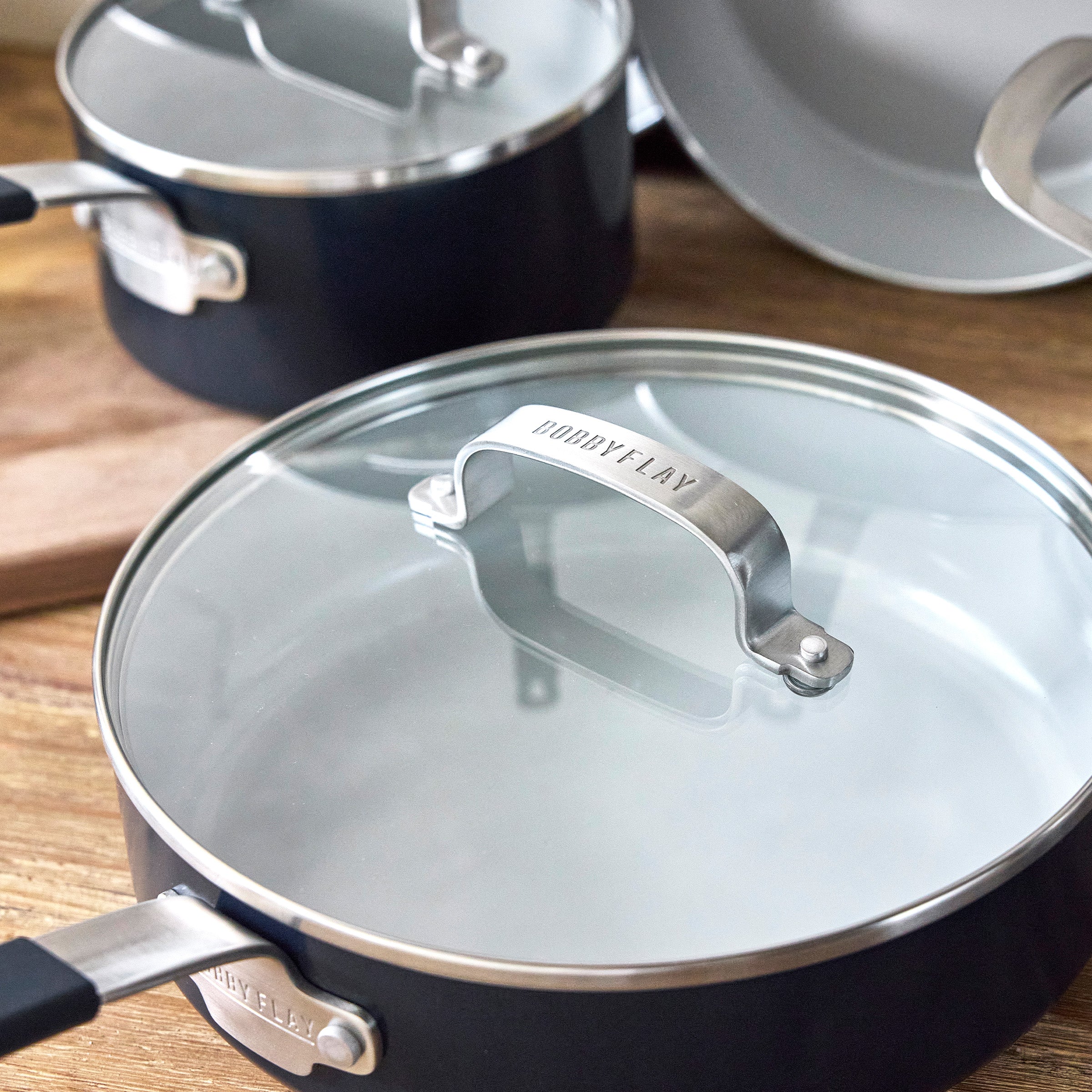 Bobby Flay standing beside GreenPan Oxford Blue aluminum cookware with engraved handles on a wooden surface.