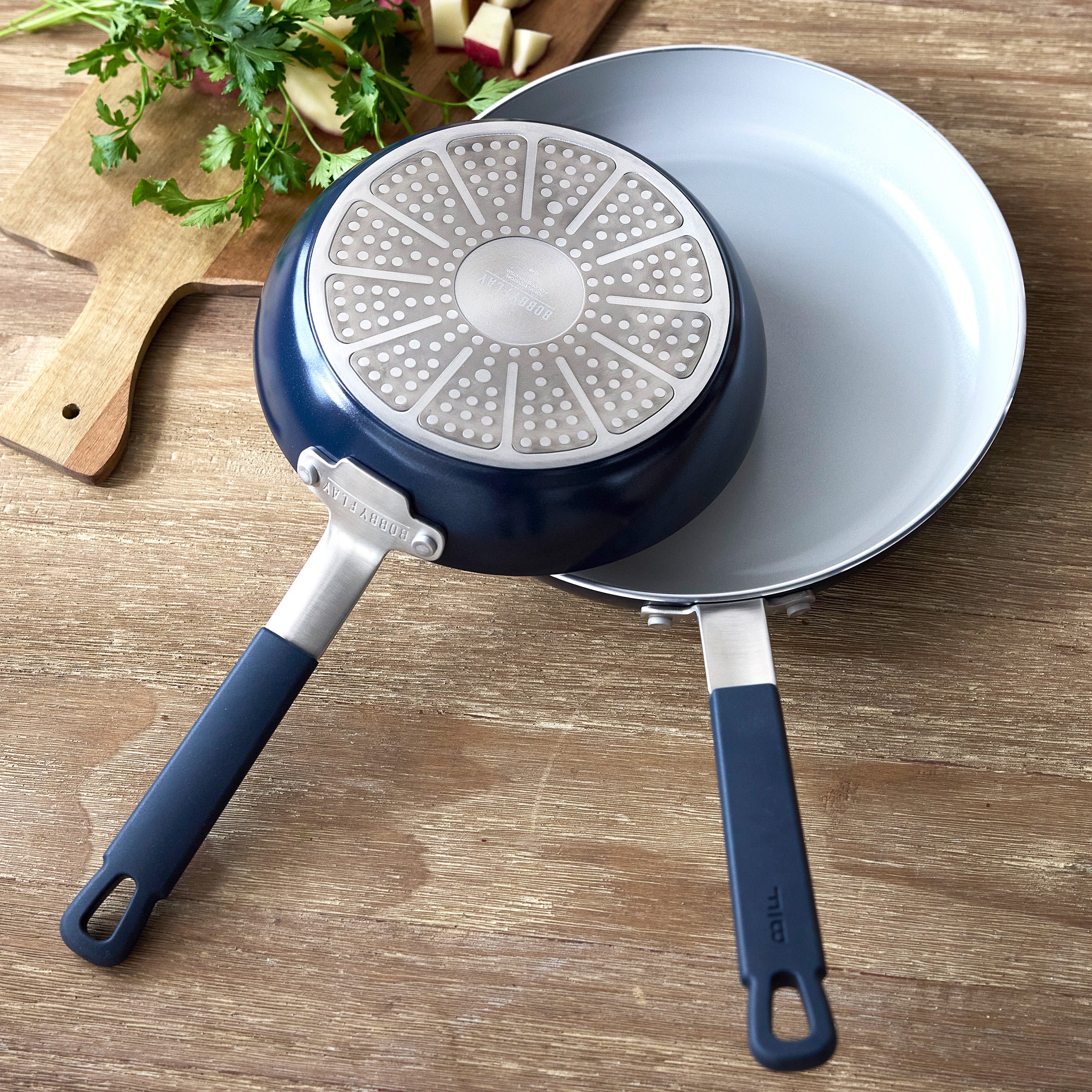 Two Oxford Blue GreenPan frying pans from a 16-piece set on a table beside a cutting board with assorted fresh vegetables.