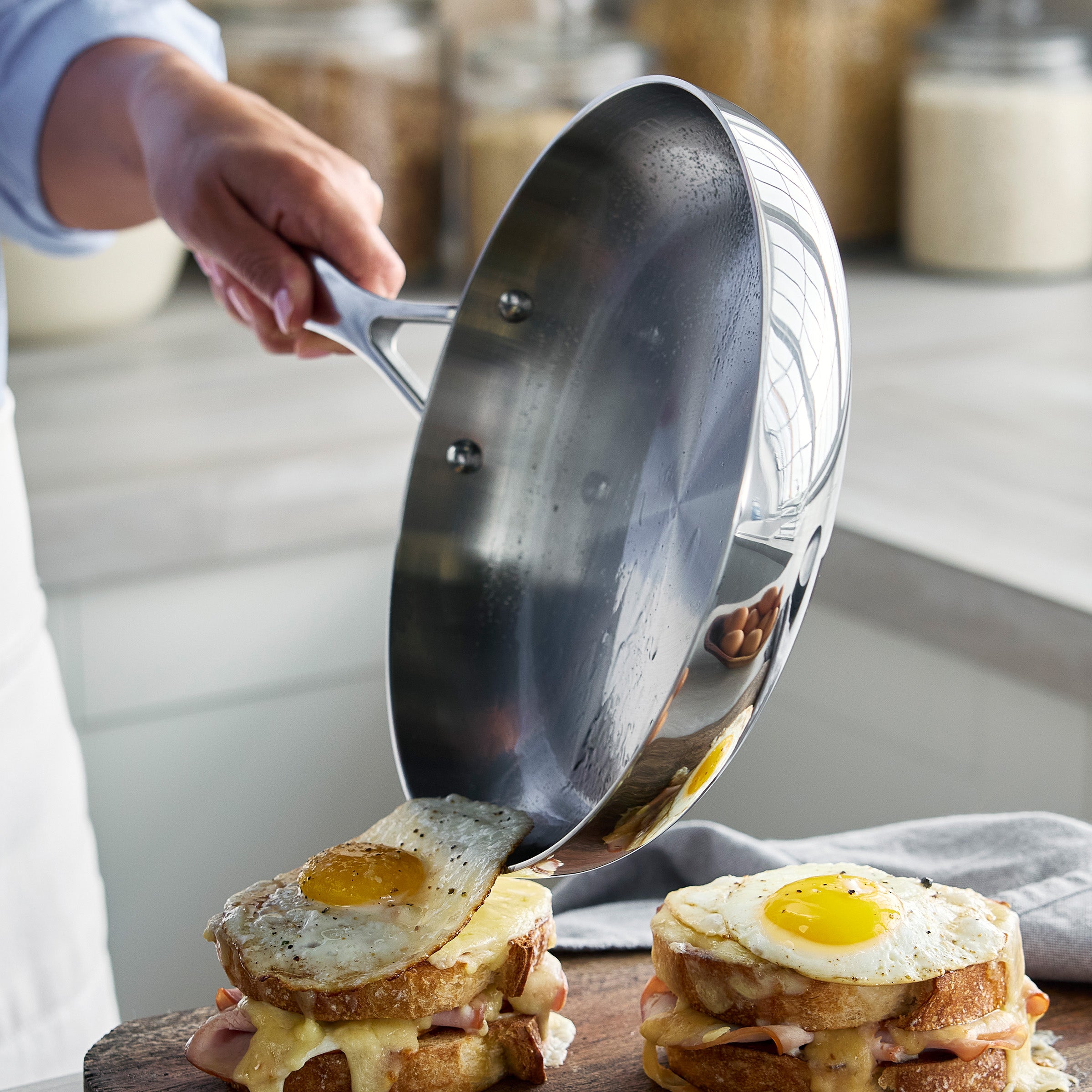 Person pouring a fried egg onto a stack of toast using a stainless steel frying pan.