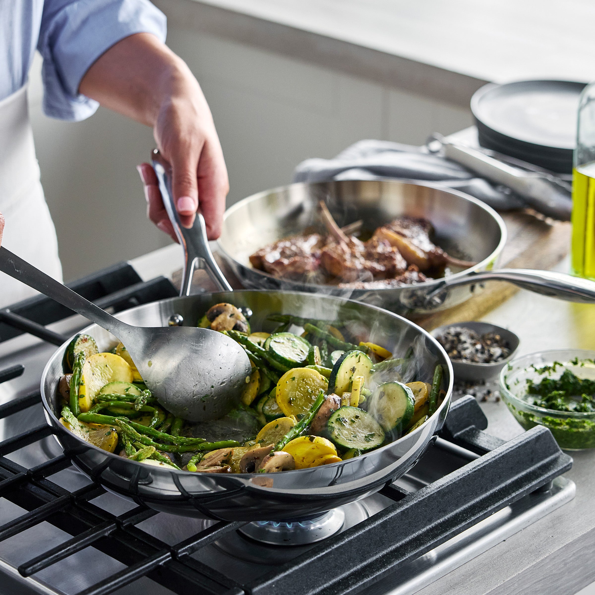 Person cooking vegetables in a frypan on a stove with another pan of food in the background.