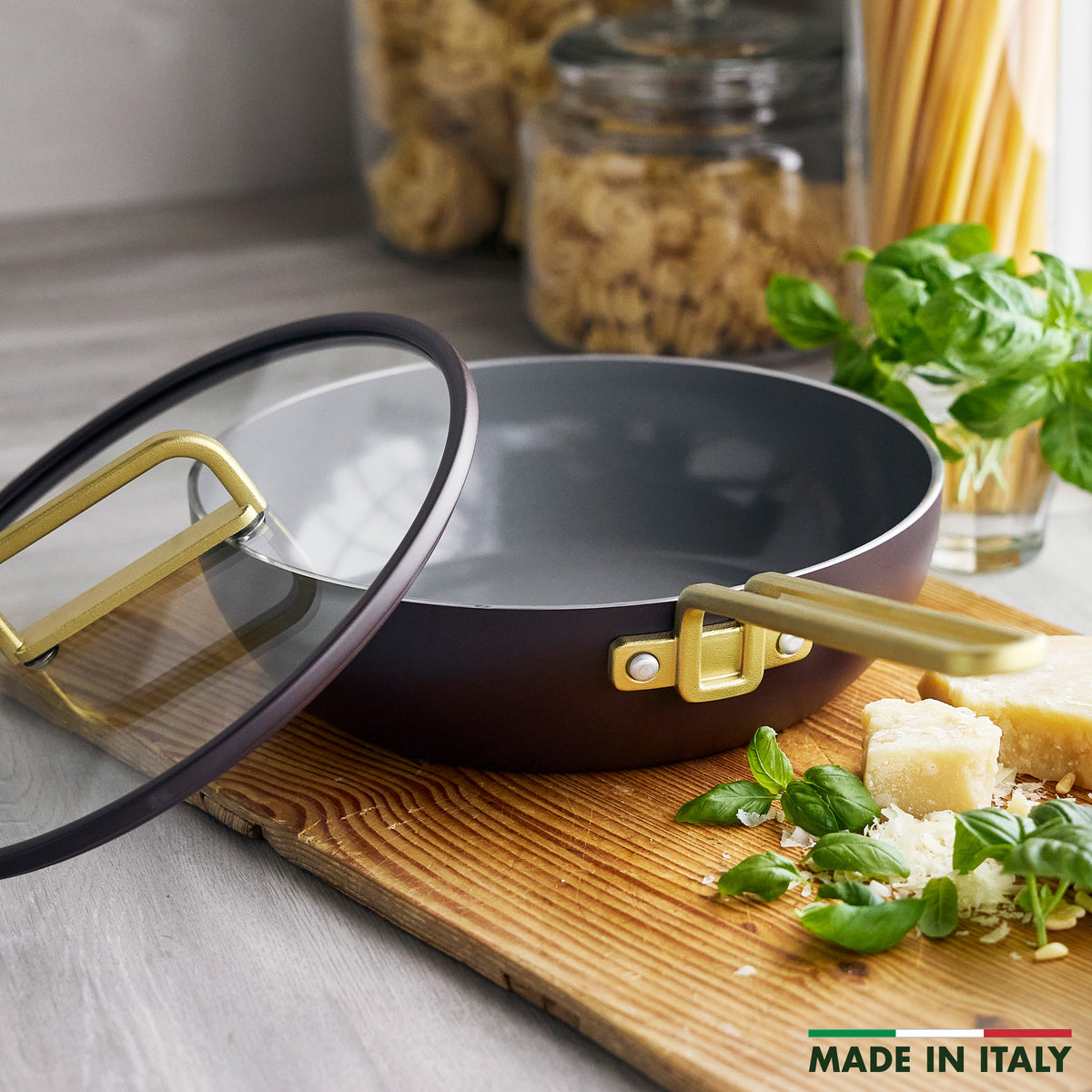 Stanley Tucci 3.25qt espresso Piccolo pan with gold handles and glass lid, displayed with basil and parmesan on a wooden board.