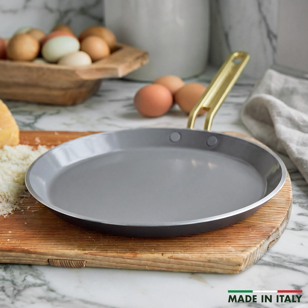 Stanley Tucci 10-inch espresso ceramic nonstick crepe and omelette pan with a gold-tone handle on a wooden cutting board.