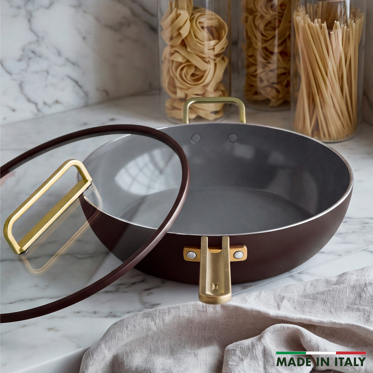 Espresso Stanley Tucci 6.5-quart ceramic nonstick essential pan with gold-toned handles and a glass lid on a marble countertop.