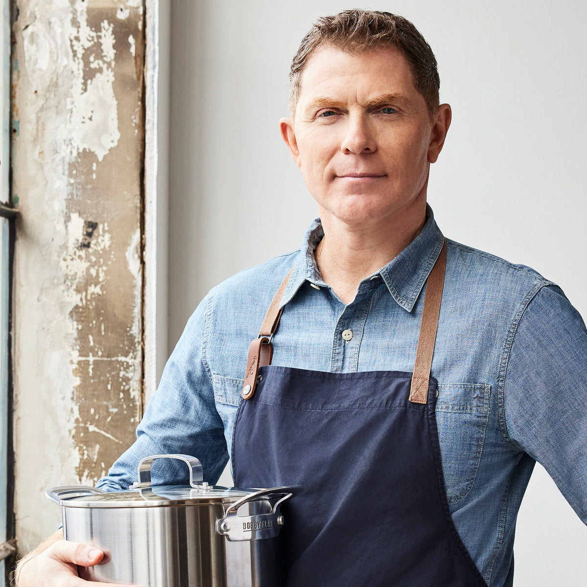 Bobby Flay by GreenPan 12-quart uncoated stainless steel stockpot with lid held by a man wearing a denim shirt and apron.