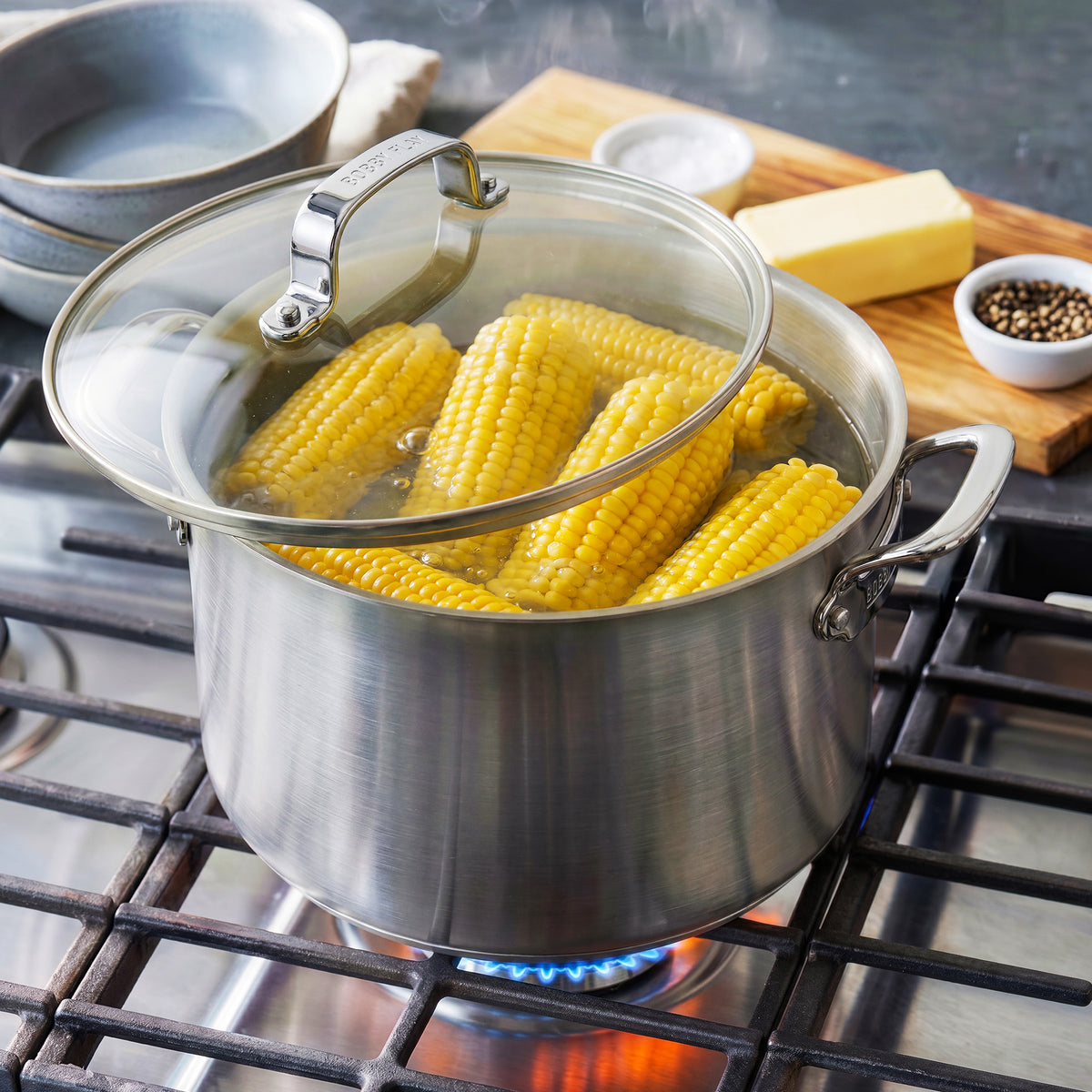 Bobby Flay by GreenPan 16-quart stainless steel stockpot with lid, filled with boiling corn on the cob on a stovetop.