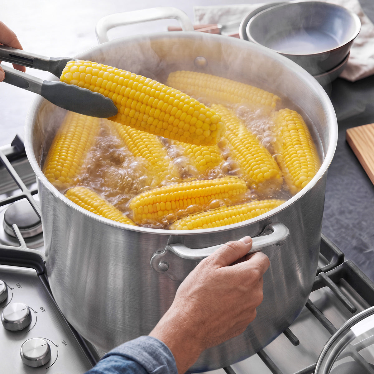 Tongs lifting an ear of corn from a Bobby Flay by GreenPan 16-quart stainless steel stockpot filled with boiling water.