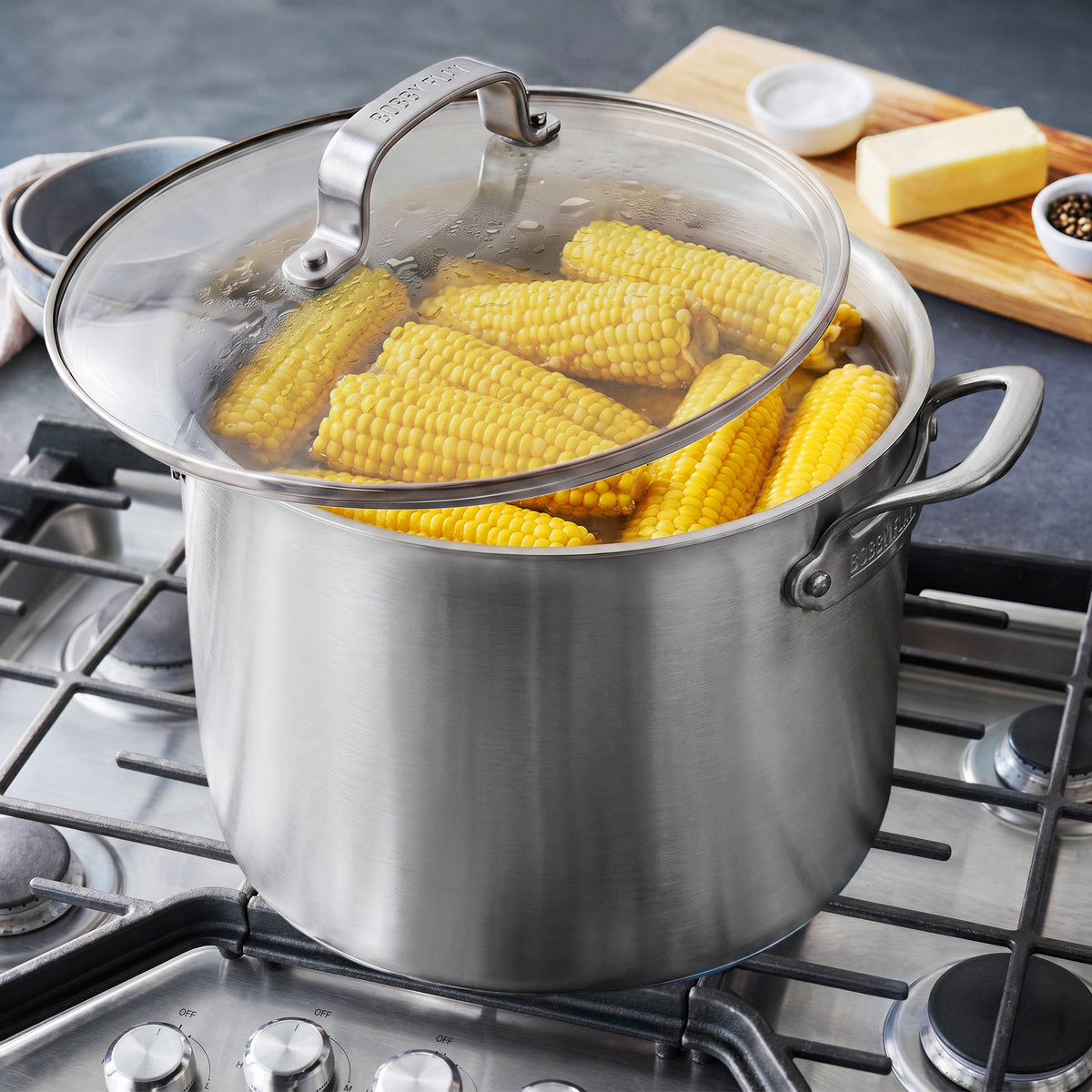 Bobby Flay by GreenPan 16-quart stainless steel stockpot with lid, filled with corn on the cob on a gas stovetop.