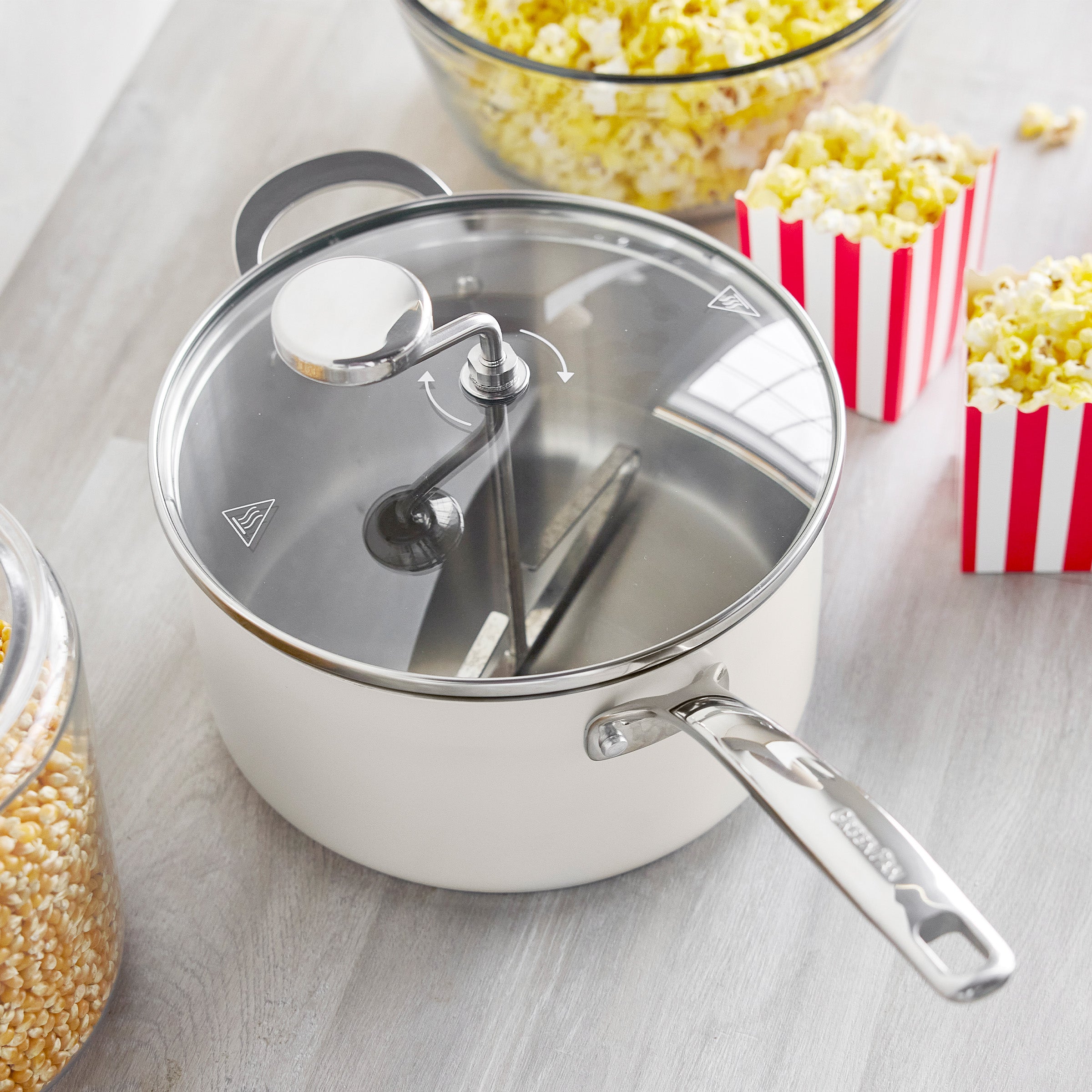Cream popcorn maker with metal handle and clear lid, surrounded by popped corn and red-striped containers on a light surface.