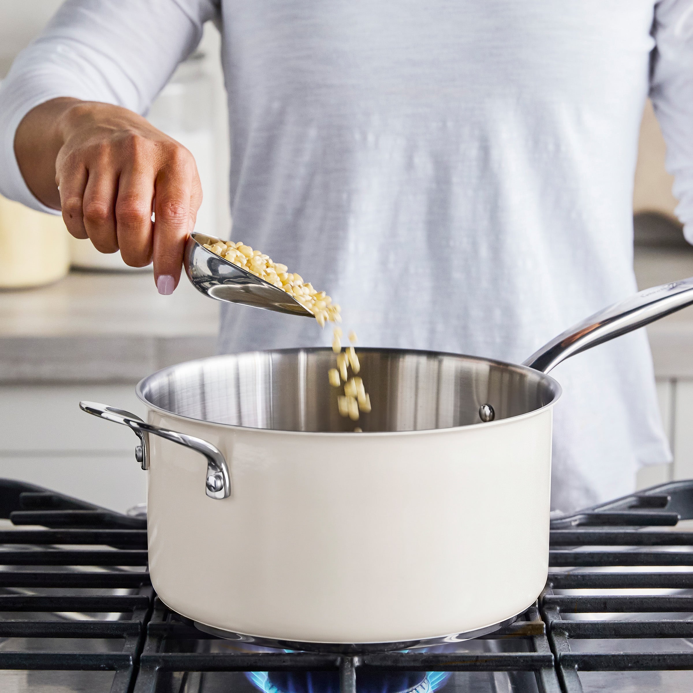 Corn kernels being poured into a Big Popper popcorn maker; cream heating in a saucepan on a blue gas flame stove.
