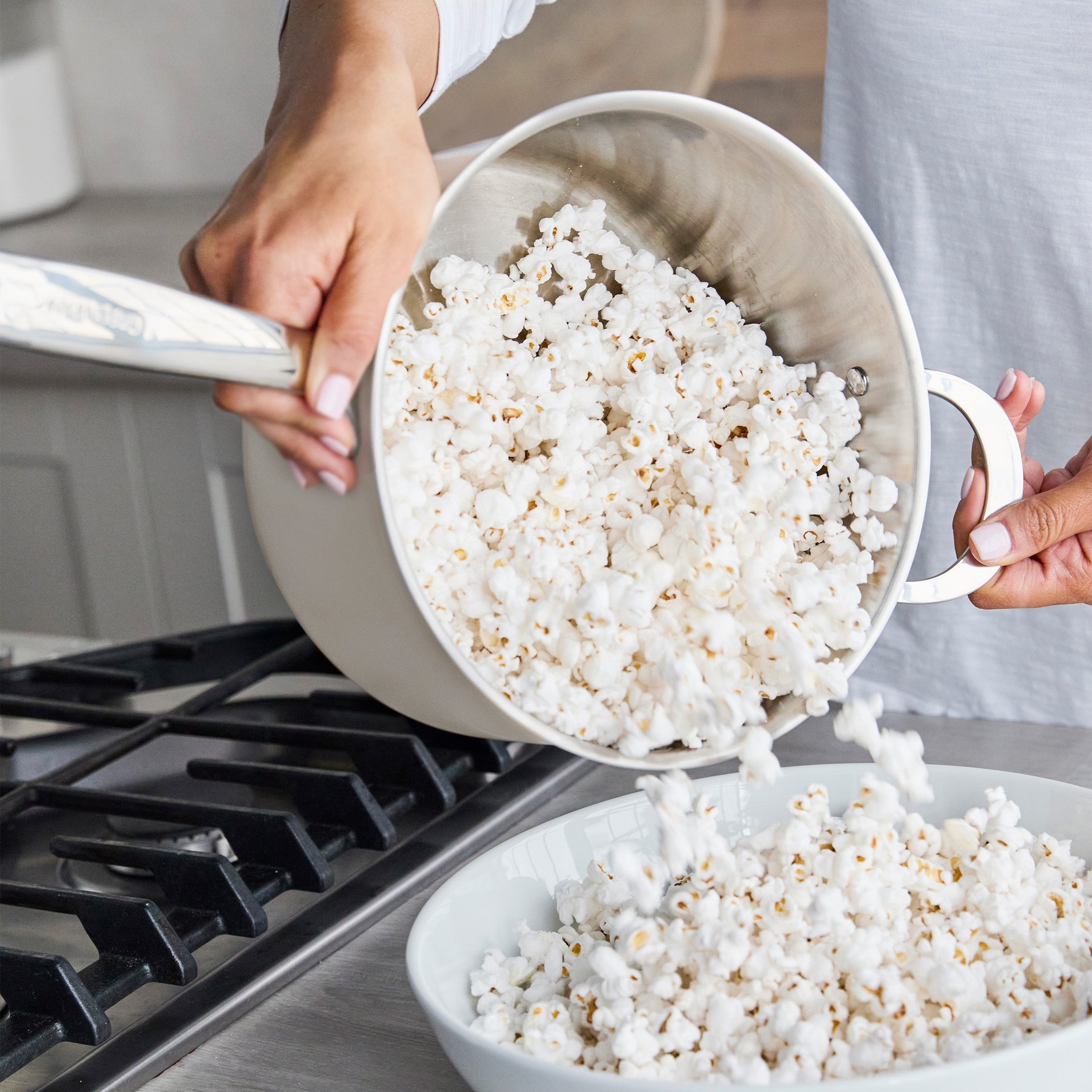 Popcorn being poured from a cream Big Popper Popcorn Maker into a white bowl on a kitchen counter near the stove.