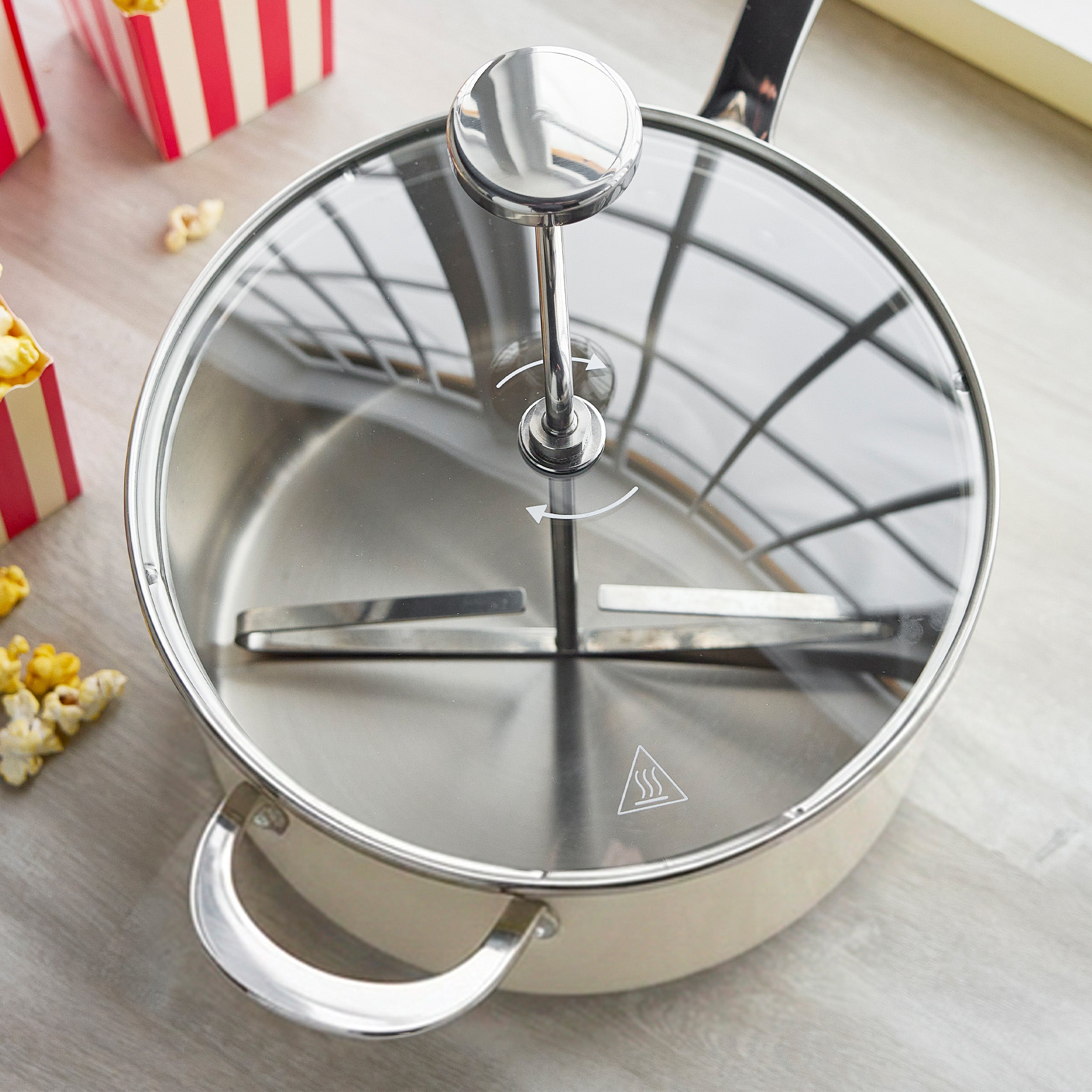 Cream stainless steel popcorn maker with glass lid, metal stirring rod, and popped popcorn on a light surface.