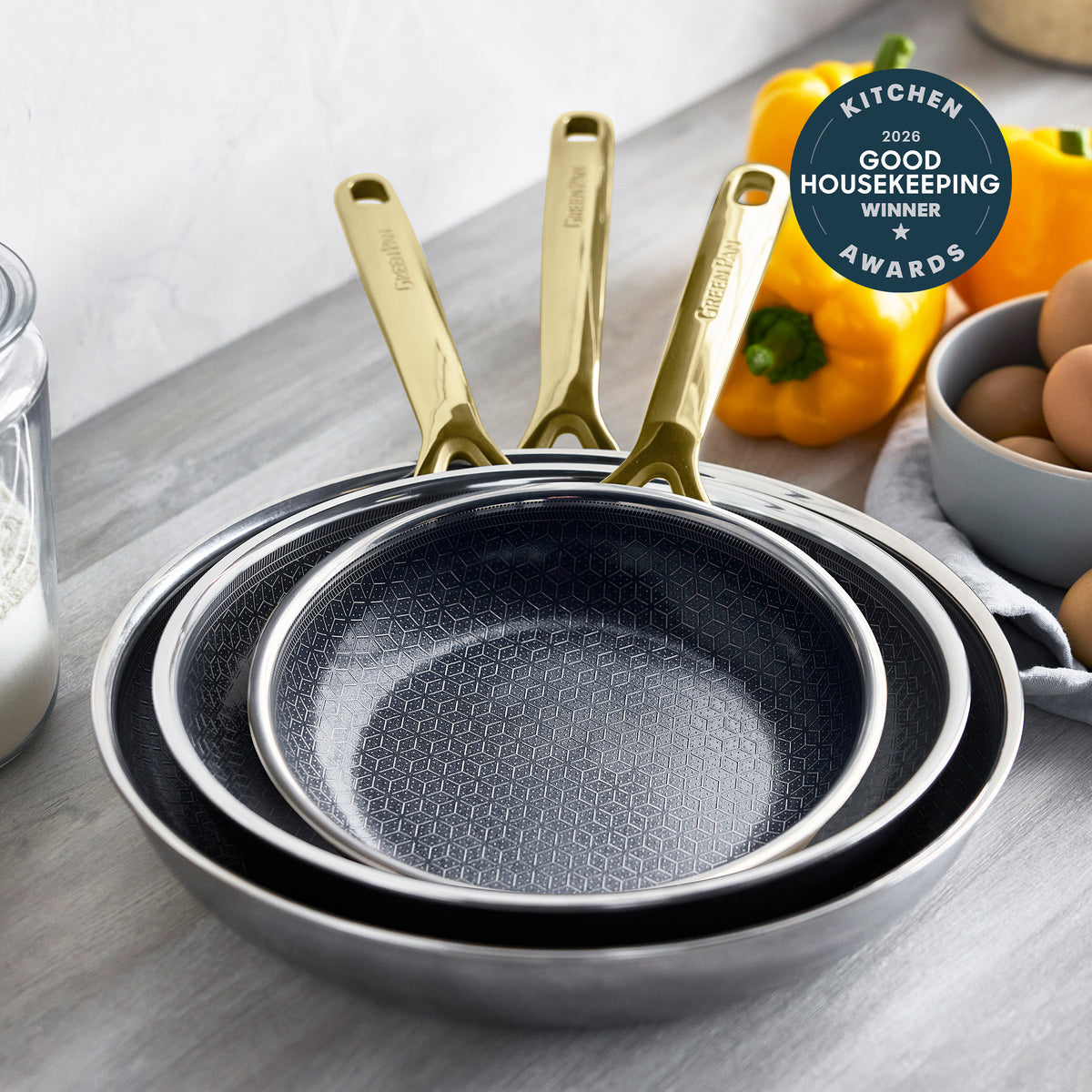 Set of three gold ceramic nonstick pans stacked on a counter with peppers, eggs, and an award badge nearby.