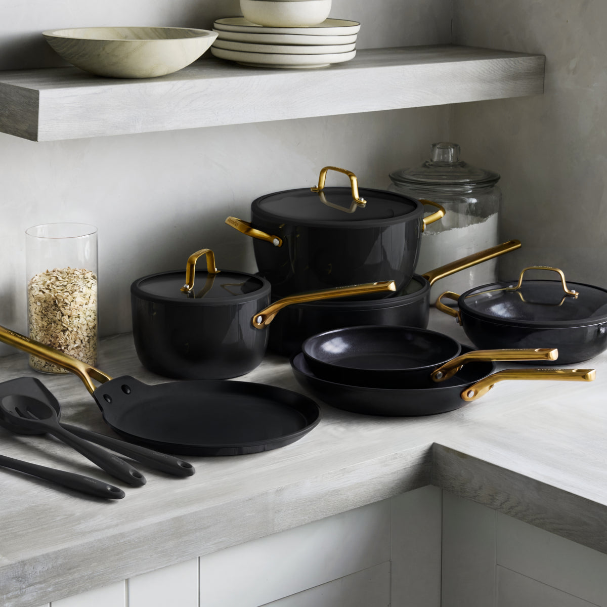 Black ceramic 14-piece cookware set with gold handles on a kitchen counter near utensils and jars.