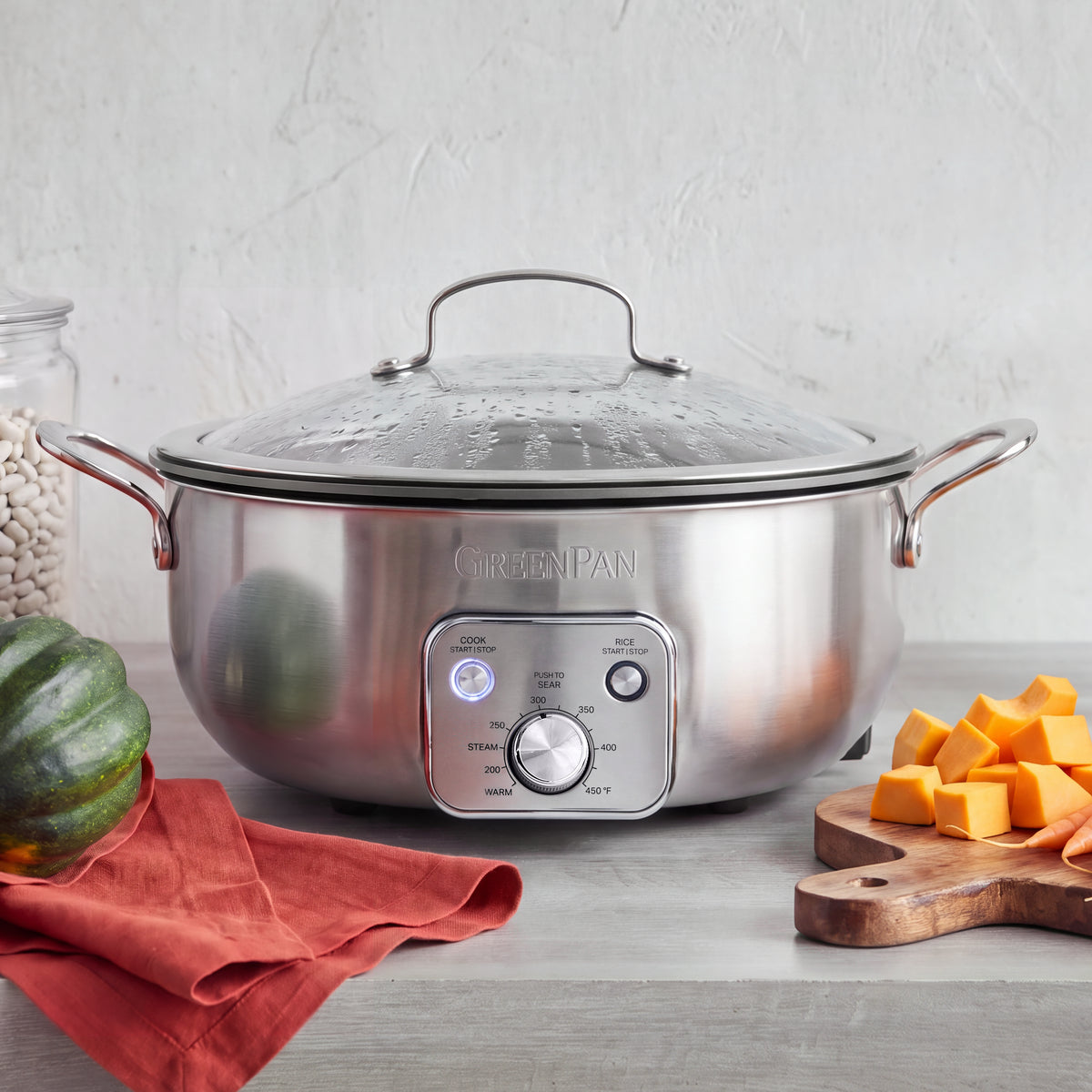 Electric wok with a glass lid sits on a kitchen counter, surrounded by assorted fresh vegetables.