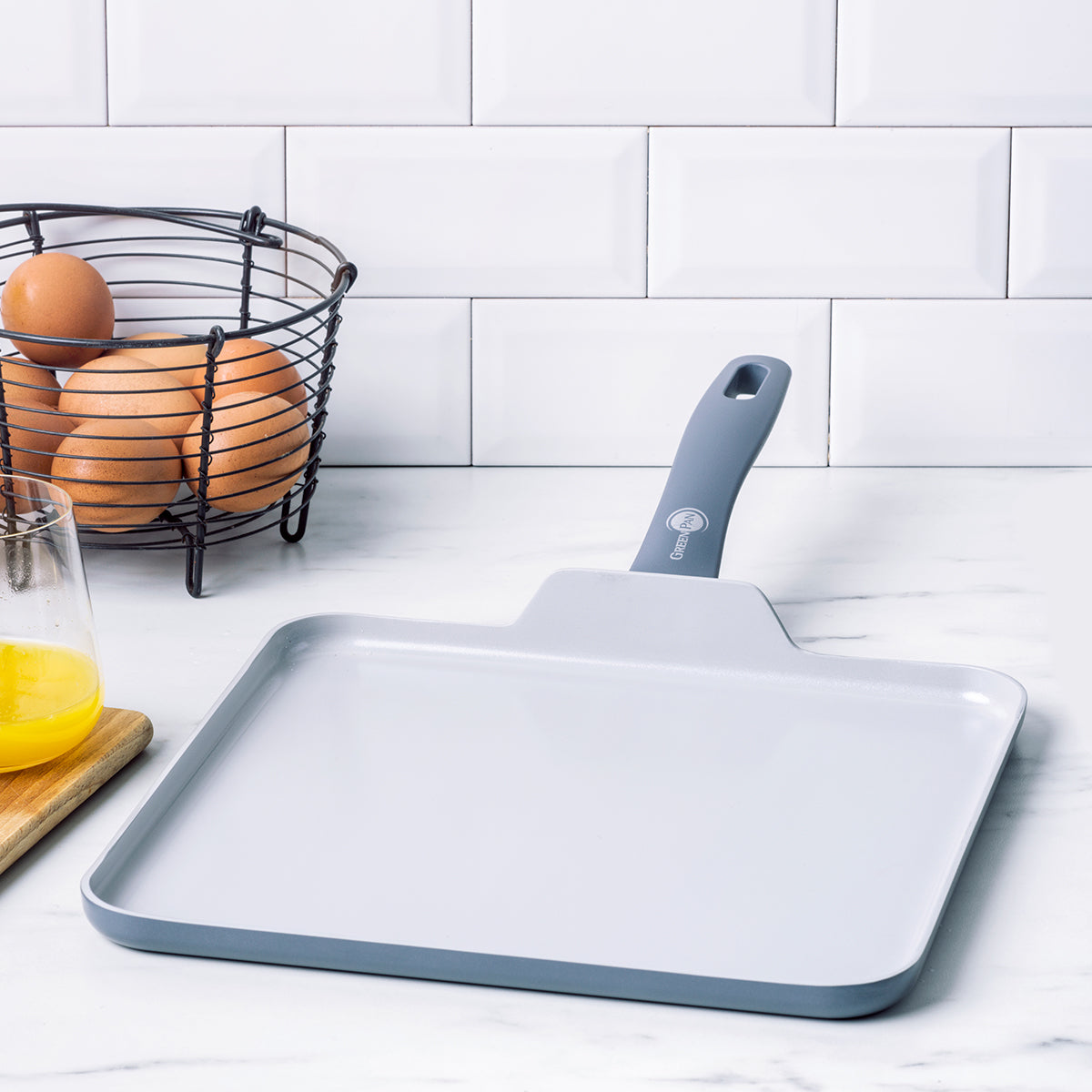 Square ceramic nonstick griddle on a white counter with a basket of eggs and a glass of orange juice nearby.