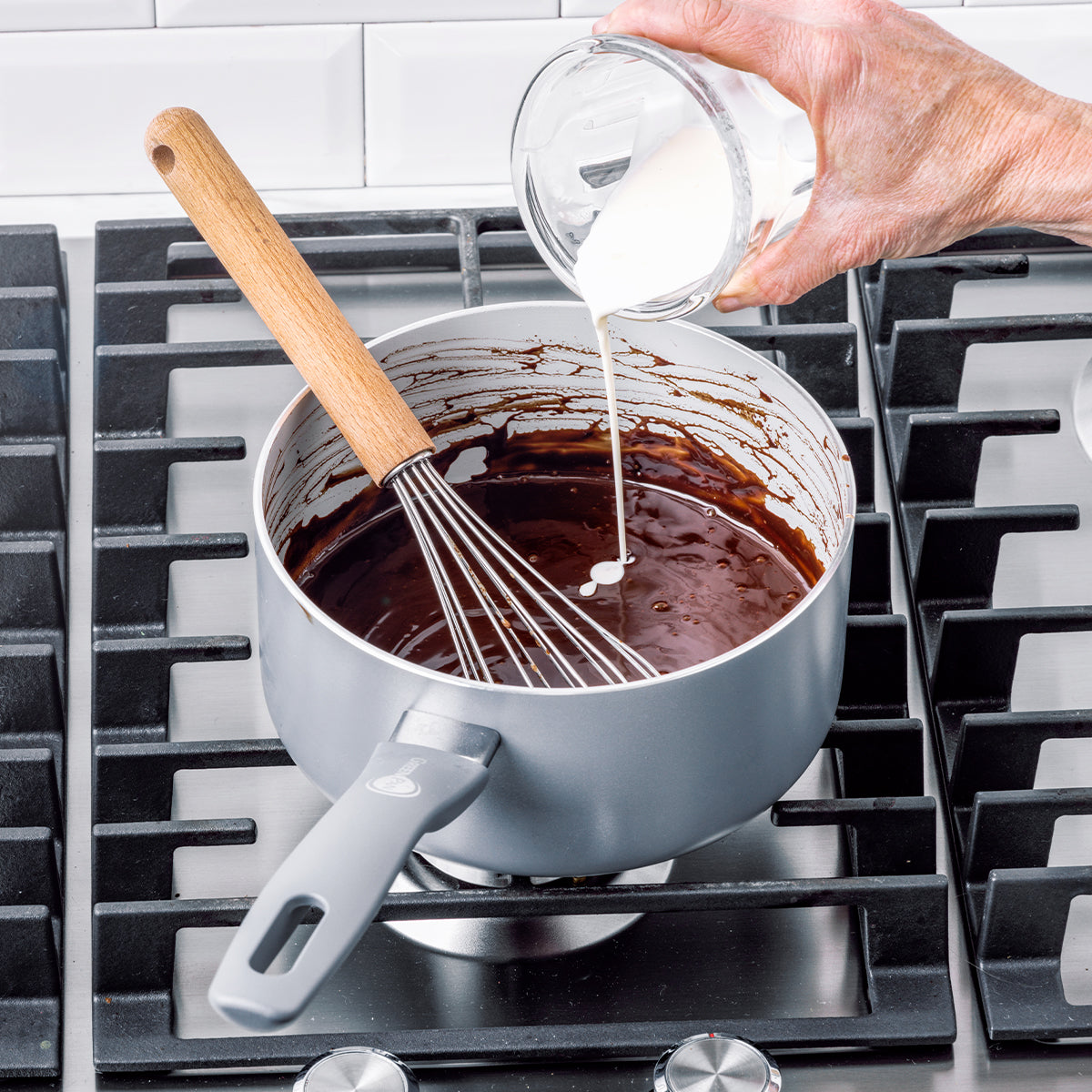 Cream being poured into a Dover ceramic nonstick saucepan with melted chocolate, whisk inside, on a stovetop.