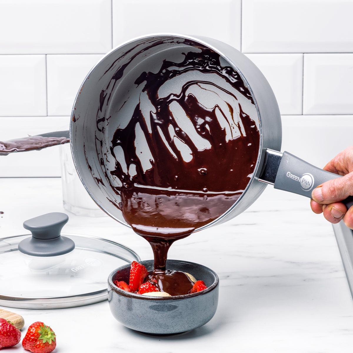 Chocolate sauce pours from a Dover ceramic nonstick saucepan onto strawberries in a bowl on a white counter.