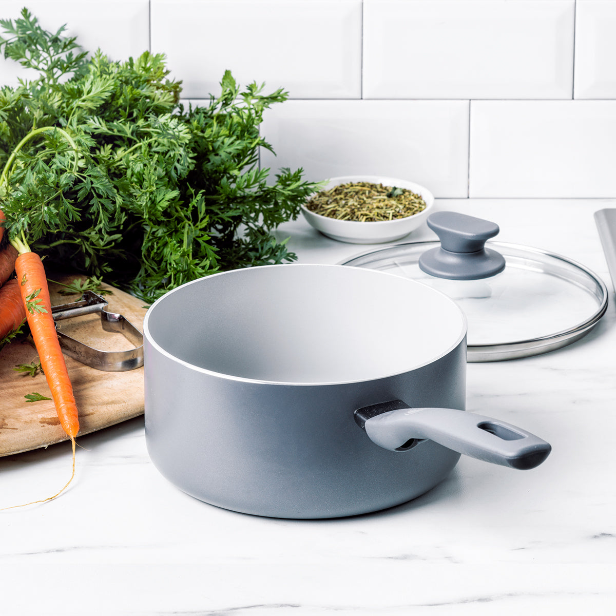 Ceramic nonstick saucepan with lid on countertop next to carrots, herbs, and a wooden cutting board.