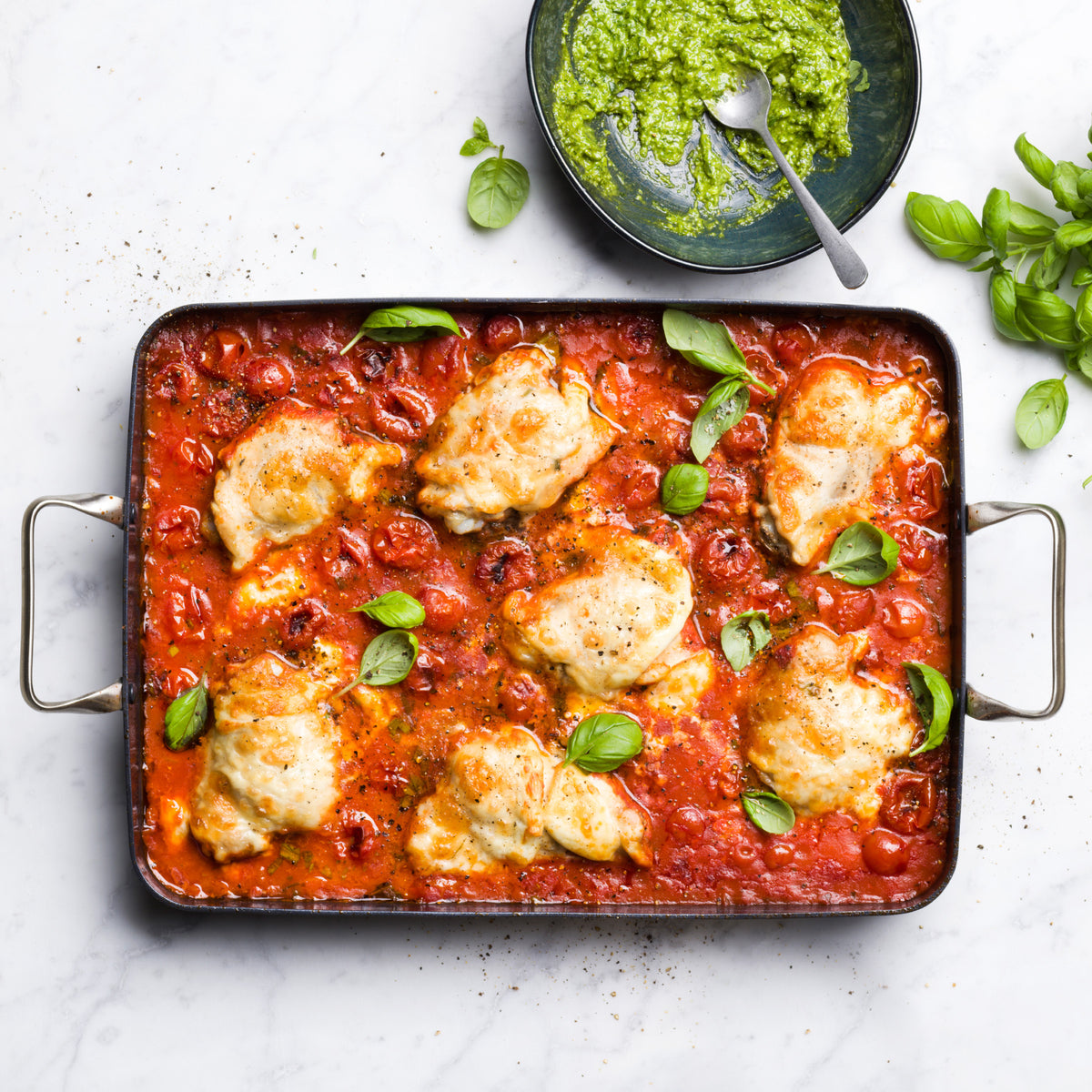 Chicken thighs in tomato sauce topped with basil in a 13.8″ x 10″ ceramic rectangular roasting pan.